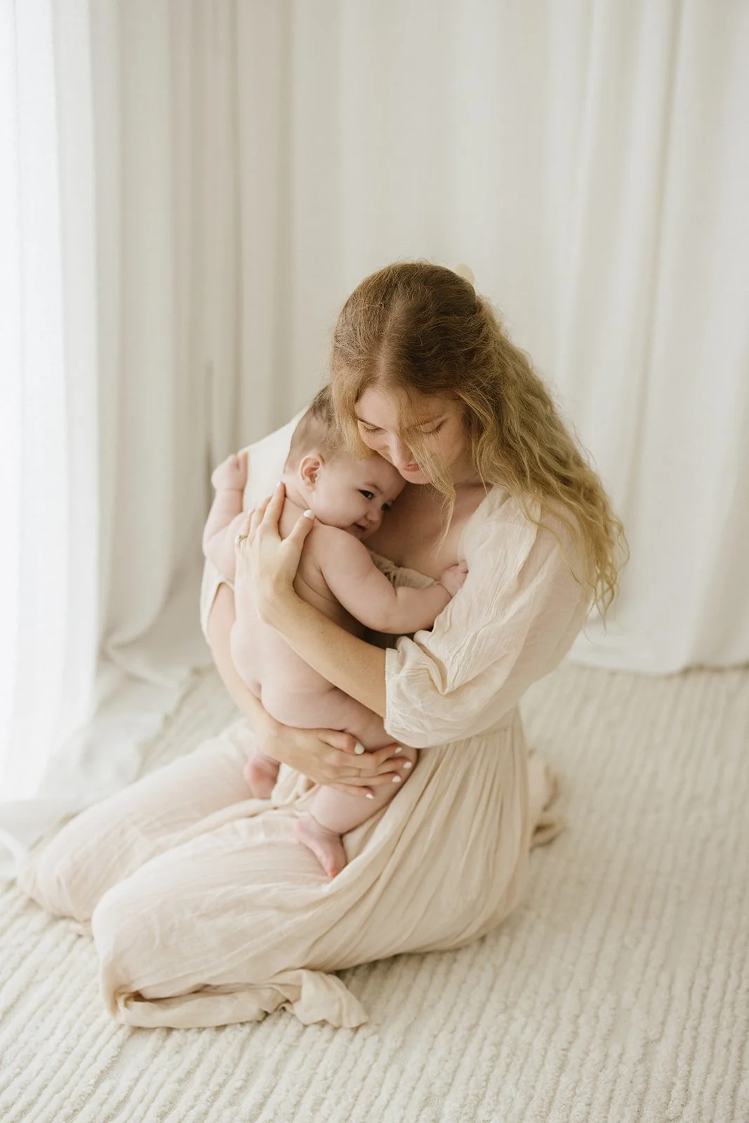 A woman with long, curly hair sitting on a beige carpet, holding her smiling baby close to her chest near a white curtain background.