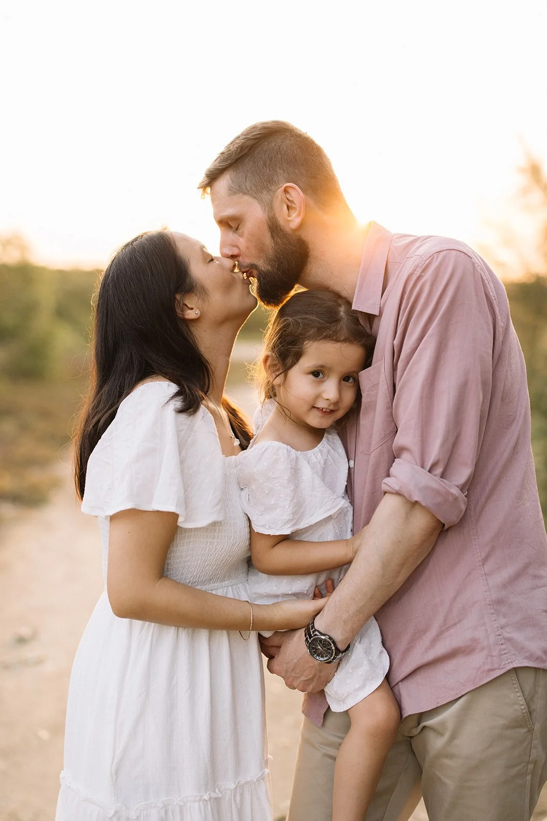 A family of three, a man, woman, and young girl, sharing a kiss outdoors at sunset. Taken by Wylde Folk Studio, Brisbane. Family photographer.