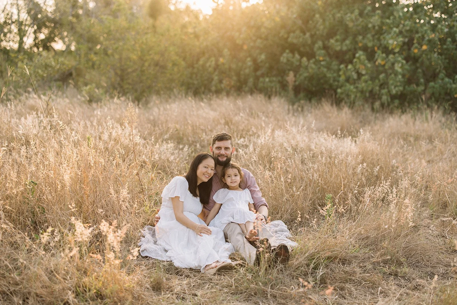 Family of three sitting together on a blanket in a field with tall grass, during sunset, with trees in the background. Taken by Wylde Folk Studio, Brisbane. Family photographer.