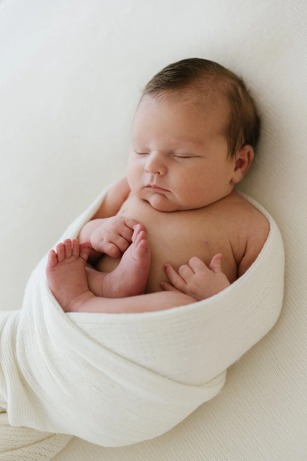 Newborn baby sleeping peacefully, wrapped in a white blanket, lying on a soft surface.