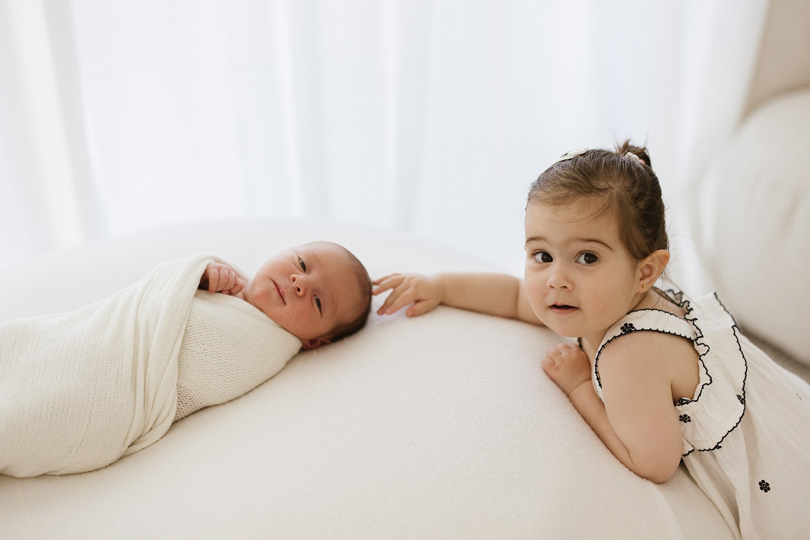 Newborn baby wrapped in white swaddle. Baby's sister touching baby's head. Both looking at camera.