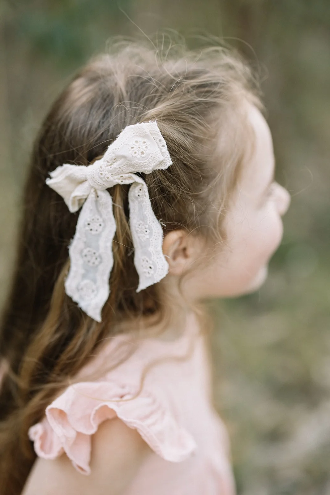 A young girl with long, wavy brown hair tied with a white lace bow, seen in profile outdoors. Taken by Wylde Folk Studio, Brisbane. Family photographer.