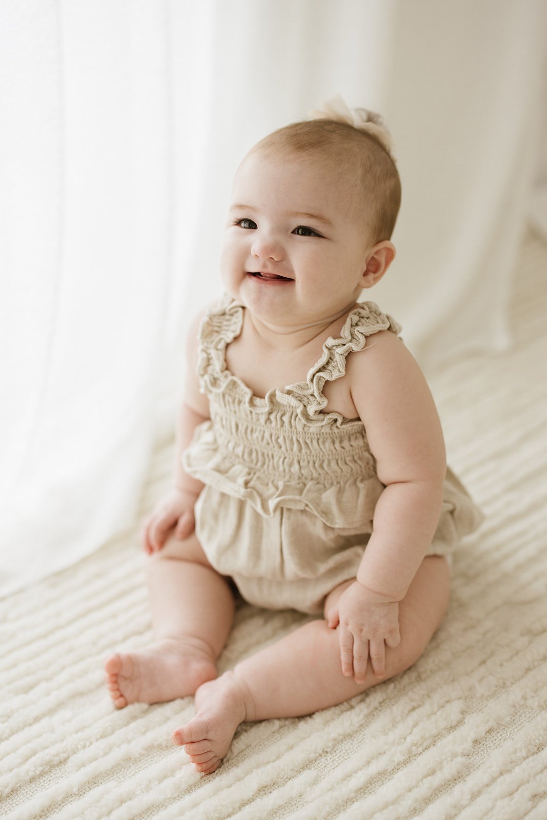 A smiling baby with light skin and short hair, sitting on a textured cream-colored blanket near a sheer curtain, wearing a beige ruffled romper.