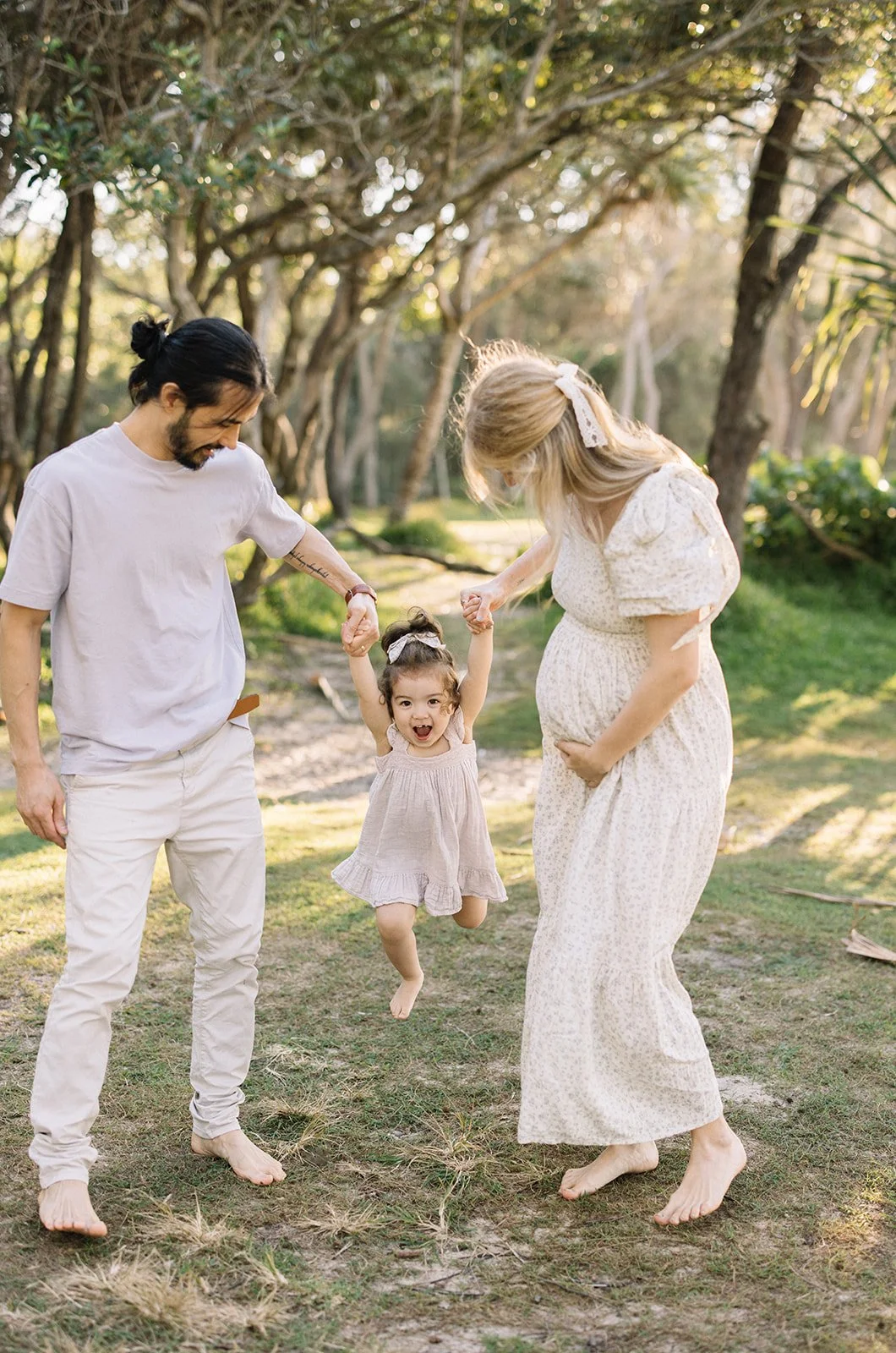 A family of three, including a man, a woman, and a young girl, holding hands and playing outdoors in a wooded park during sunlight. The girl is jumping happily, and the woman is pregnant, smiling at the girl.