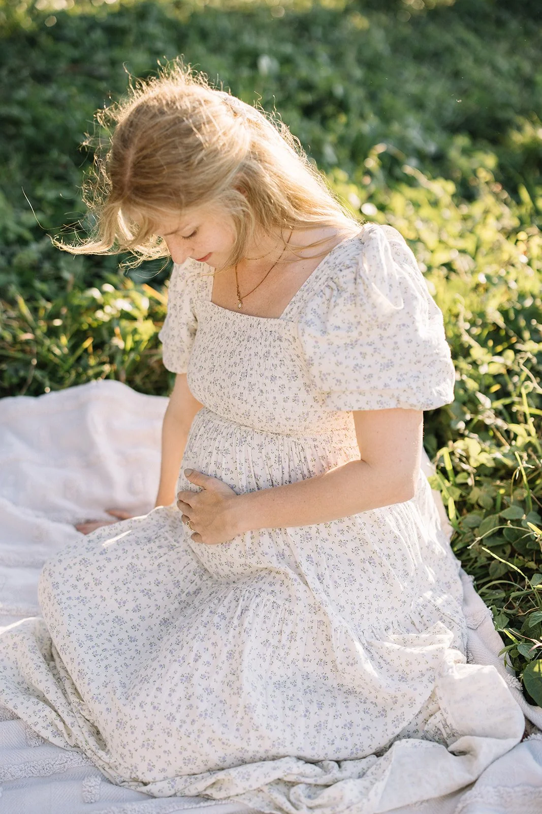 A pregnant woman in a white floral dress kneels on a white blanket outdoors, gently holding her baby bump and looking down with a serene expression, surrounded by green plants and illuminated by soft sunlight.