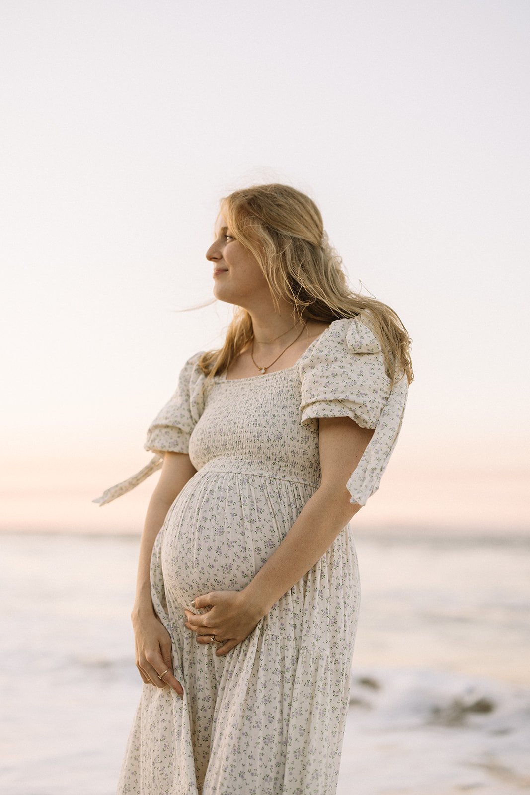 Pregnant woman in a floral dress standing on a beach at sunset, smiling and gently holding her belly. Taken by Wylde Folk Studio, Brisbane. Maternity photographer.