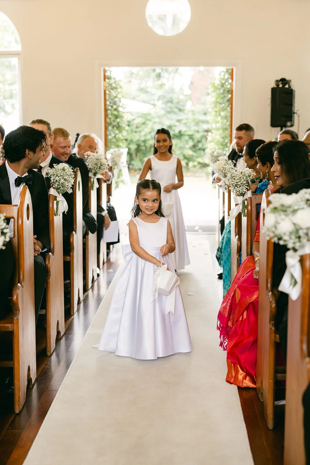 Three young girls walking down the aisle during a wedding ceremony, wearing white dresses and holding small baskets, with guests seated on either side, in a bright, sunny church setting.