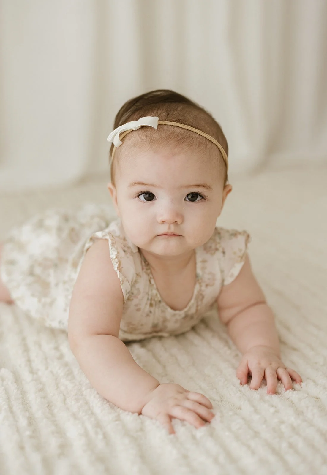 A baby girl with a cream dress and a beige headband with a white bow, lying on her stomach on a cream-colored blanket, looking directly at the camera. Taken at Wylde Folk Studio - Brisbane baby photographer.