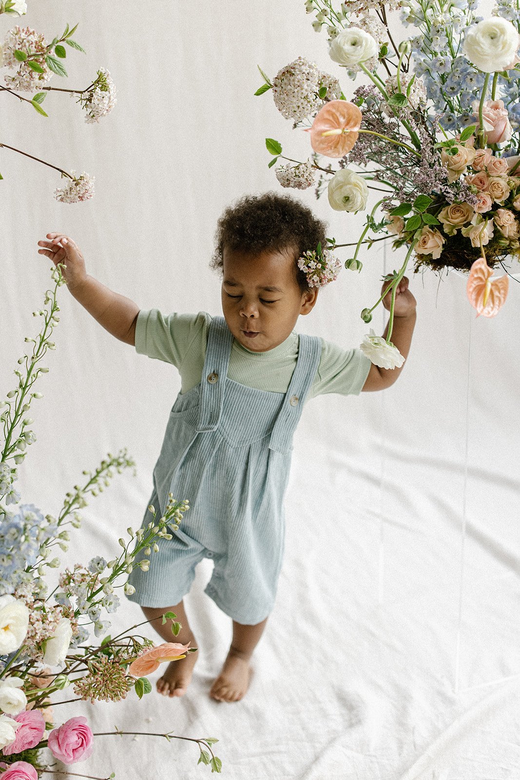 a little boy is seen dancing surrounded by flowers in a cute pastel romper for a children's clothing brand during a photoshoot