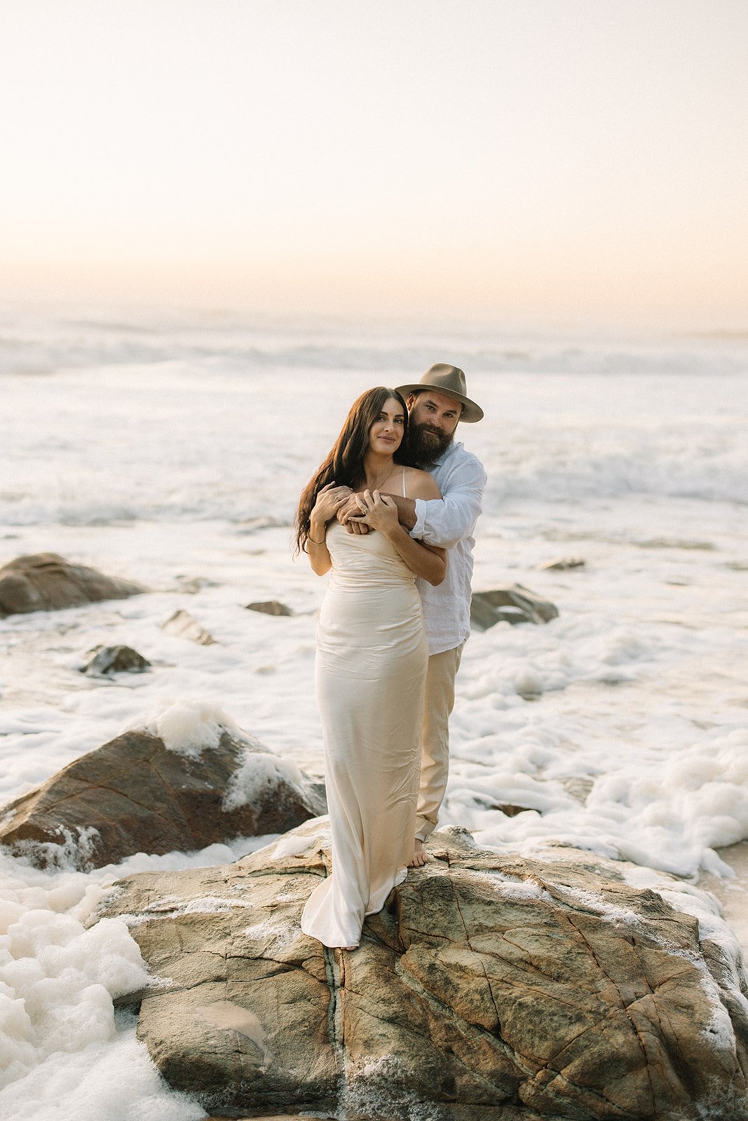 a man wraps his arms around his wife with long dark hair. They are standing on a rock at the beach and the waves are surrounding them