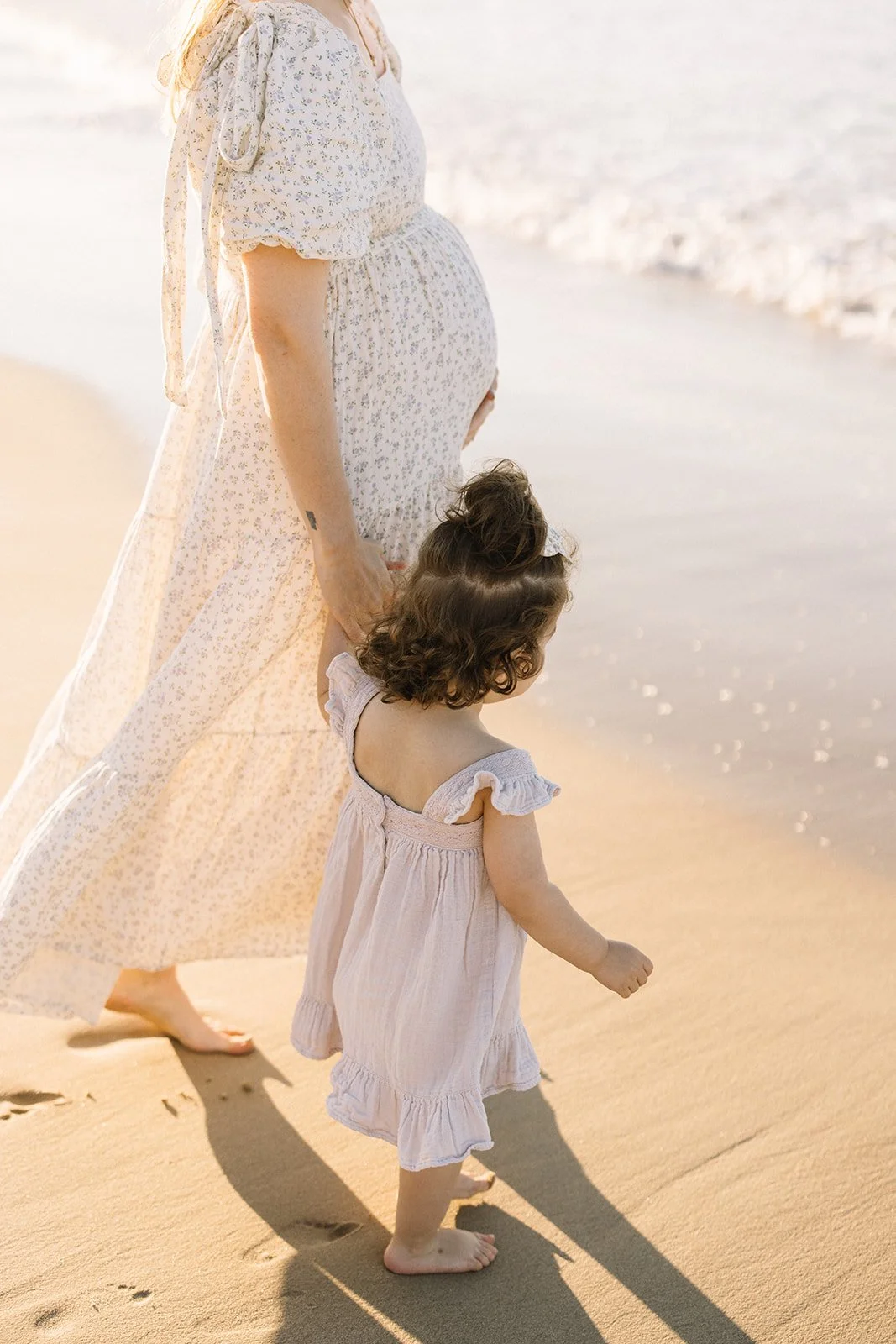 A pregnant woman and a young girl walking barefoot on the beach near the shoreline during sunset or sunrise. Taken by Wylde Folk Studio, Brisbane. Maternity photographer.