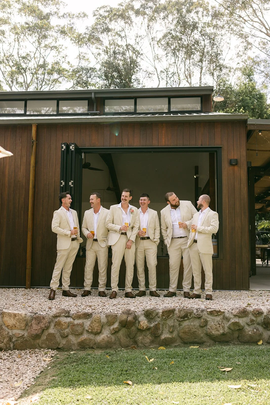 A group of seven men dressed in cream-colored suits standing outside a wooden building, holding drinks, and smiling at each other.