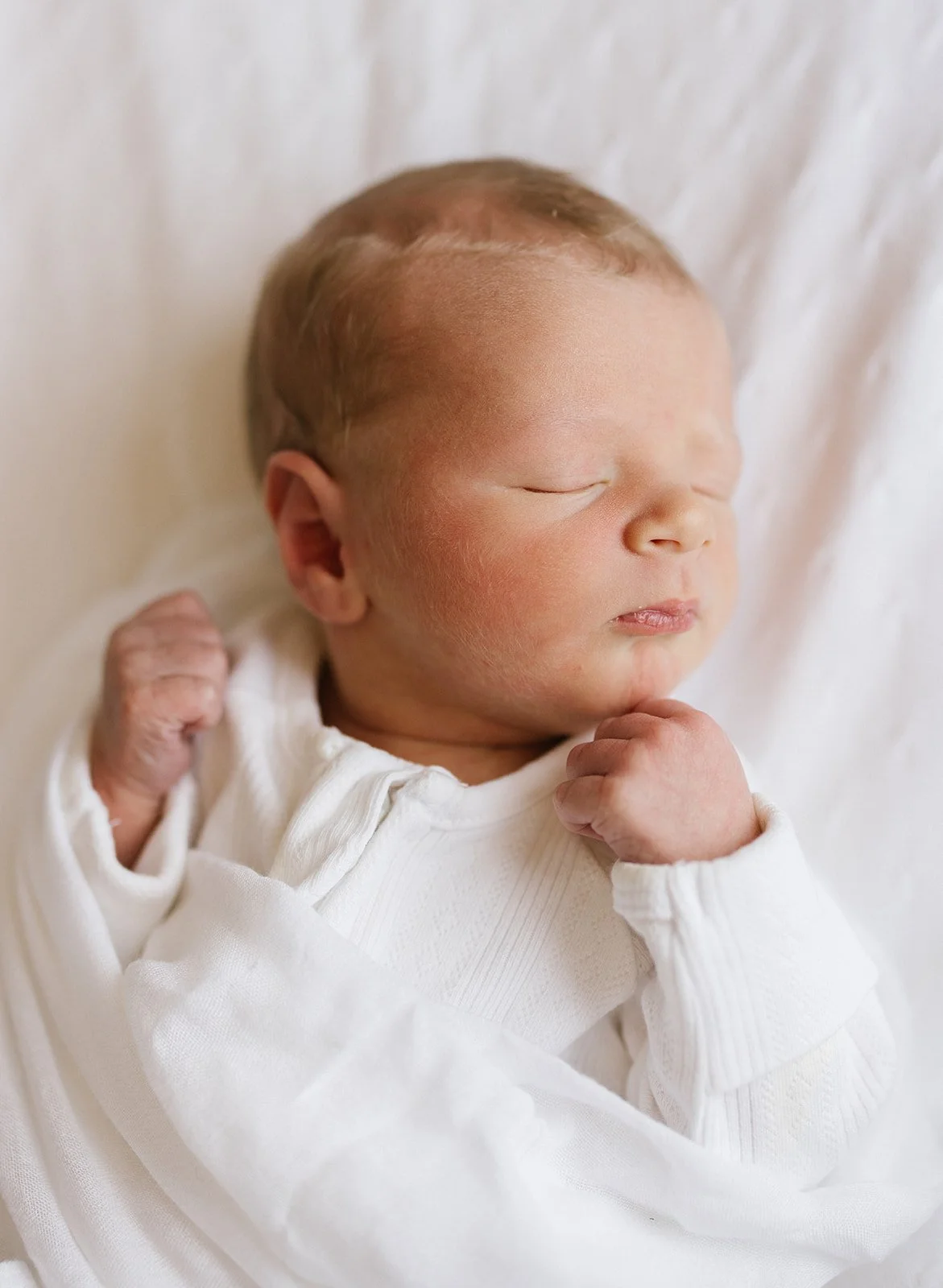 Close-up of a sleeping newborn baby with closed eyes, wearing a white outfit, resting on a white blanket.