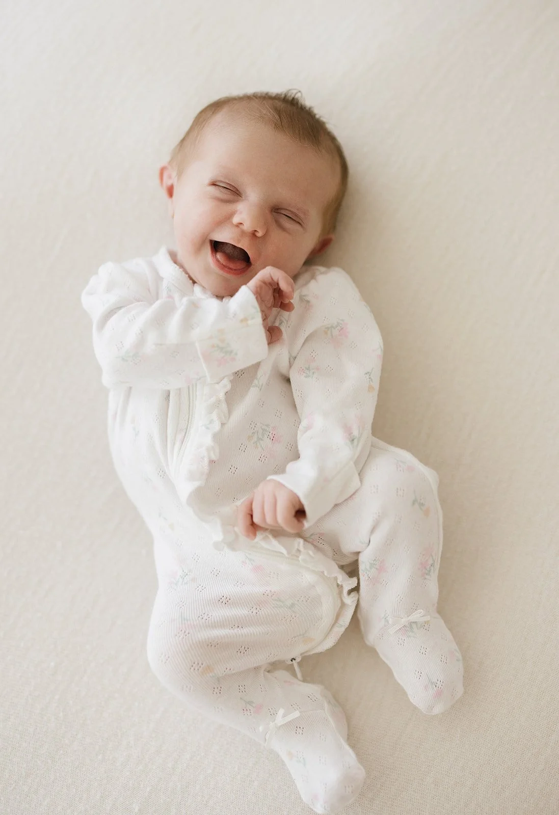 Newborn baby with light brown hair smiles with eyes closed. Lying on white beanbag and wearing a white onesie.