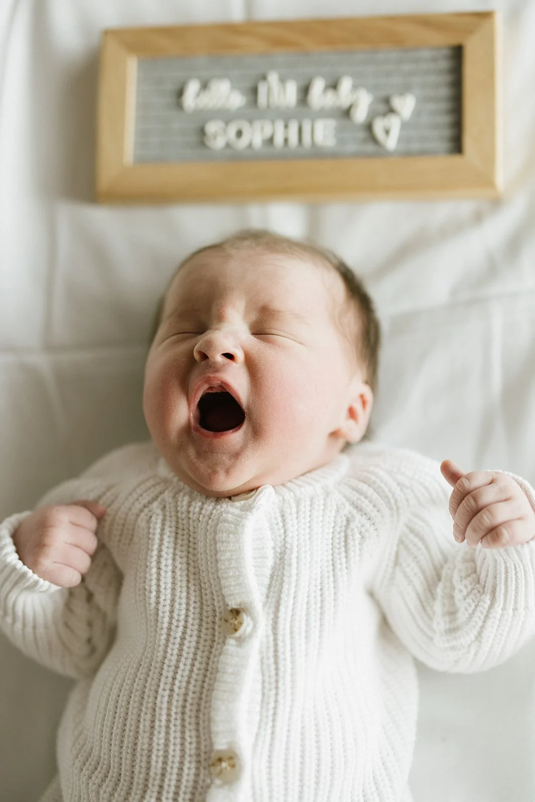 A newborn baby yawning while lying on a white blanket, wearing a cream-colored knit sweater, with a wooden framed sign above that reads 'the tiny ♥ SOPHIE' with a heart symbol.