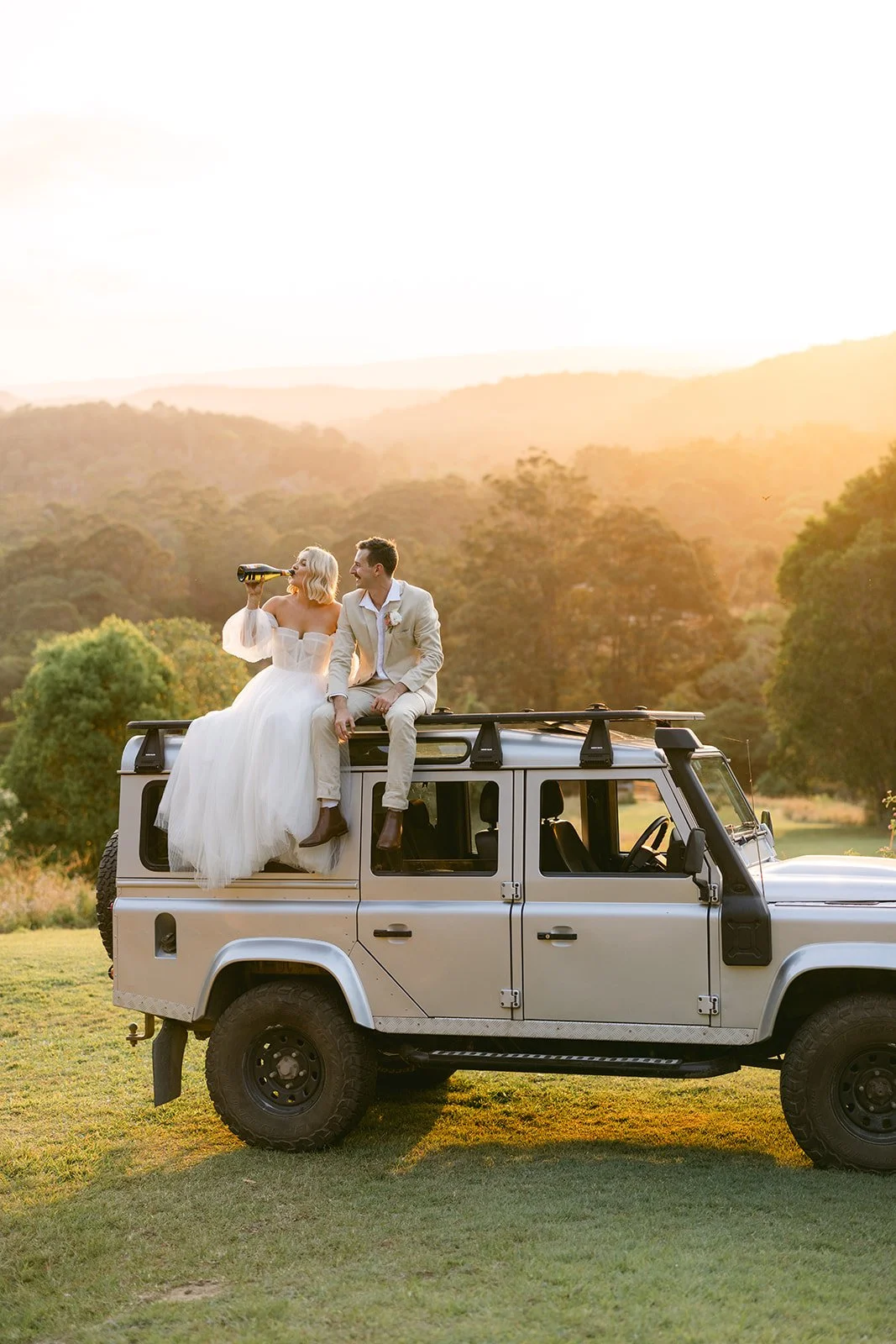 Bride and groom sitting on top of a Land Rover vehicle outdoors during sunset, with the bride drinking from a bottle and the groom smiling.
