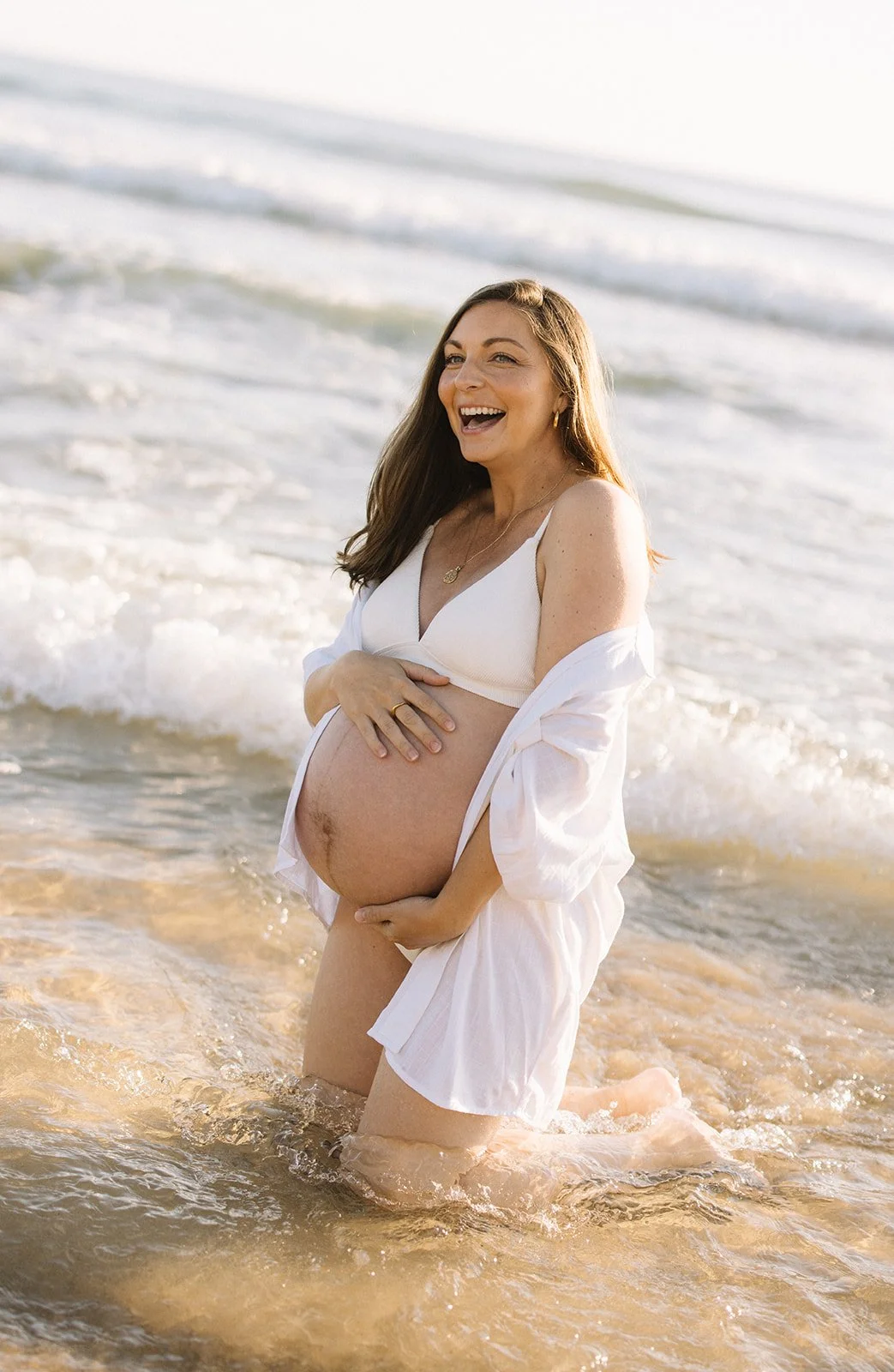 Pregnant woman in white clothing kneeling in water at the beach, smiling and holding her belly. Taken by Wylde Folk Studio, Brisbane. Maternity photographer.