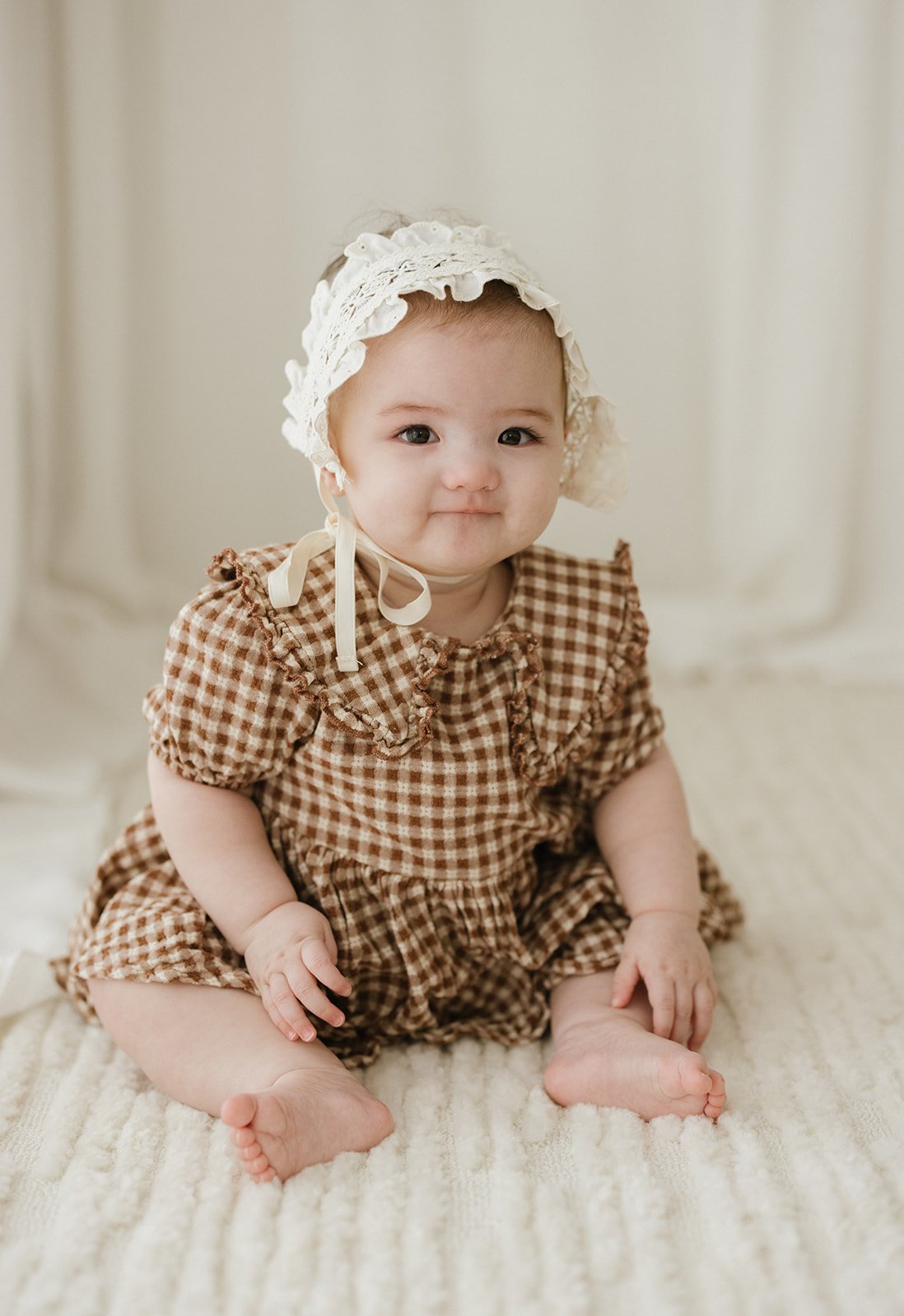 A baby girl sitting on a soft white blanket, wearing a brown checkered dress with ruffles and a white lace bonnet with a ribbon tie. Taken at Wylde Folk Studio - Brisbane baby sitter photography.