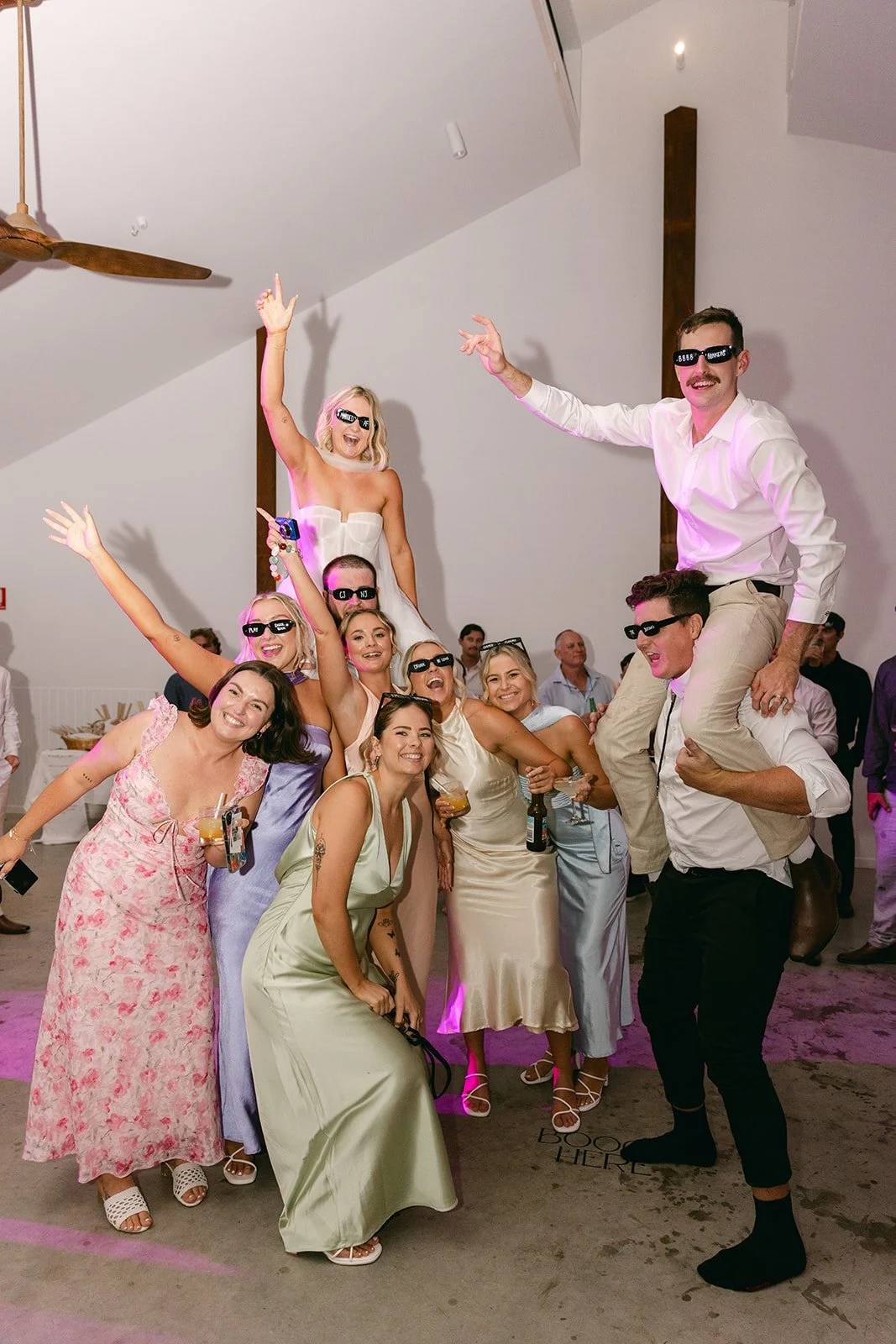 Group of people at a celebration, some wearing sunglasses, lifting and supporting a woman on their shoulders, all smiling and dressed in formal wear.
