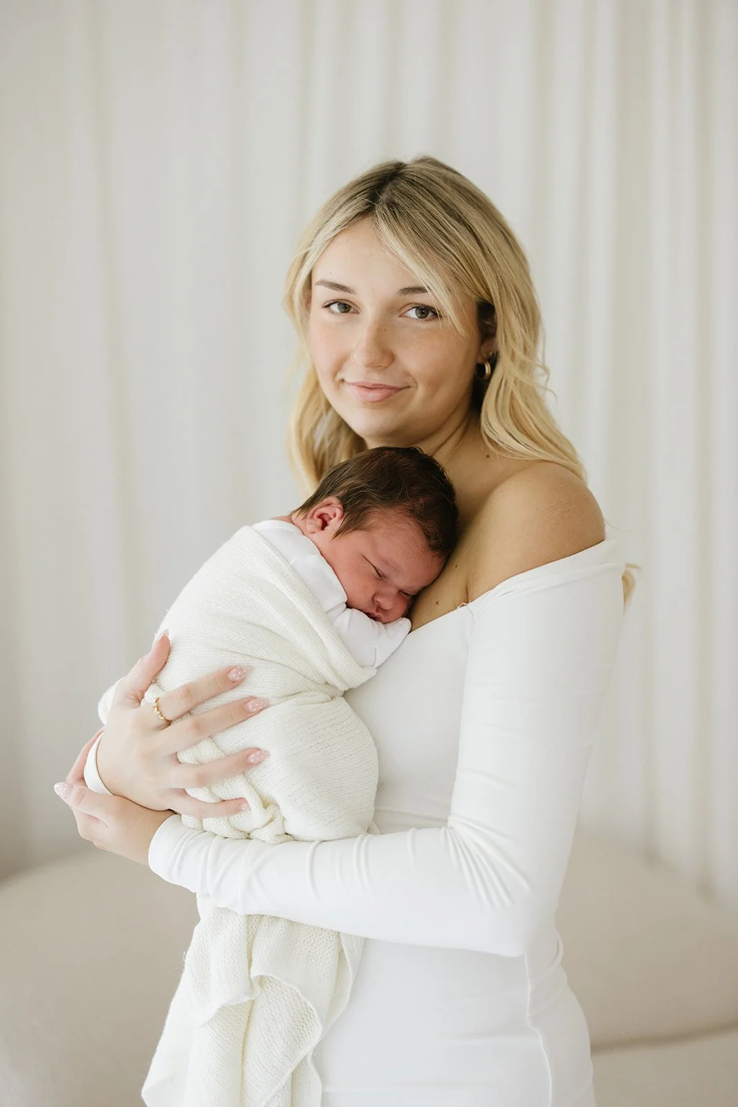 A woman with blonde hair holding a sleeping newborn baby wrapped in a cream blanket.