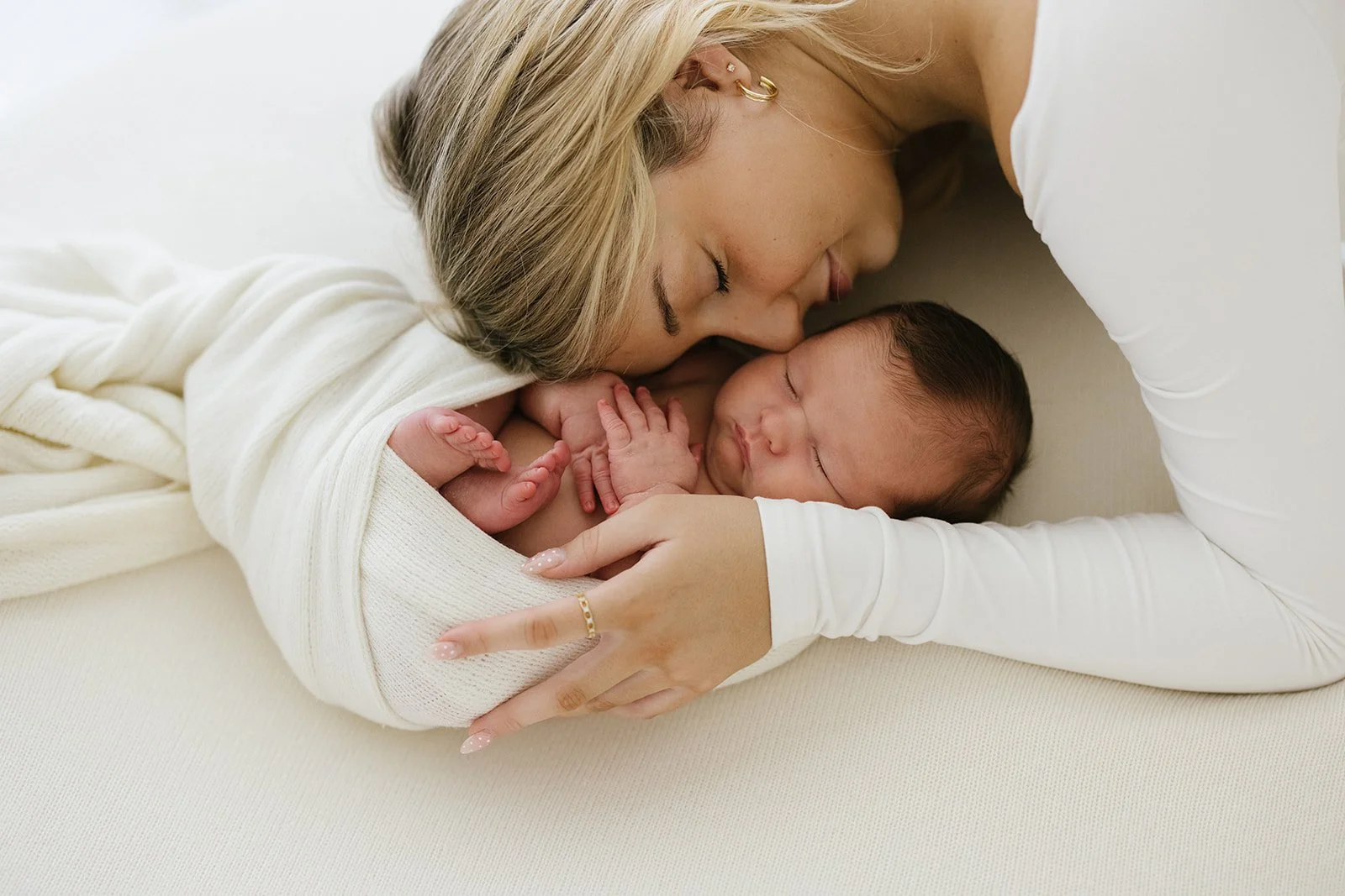A mother with blonde hair and earrings gently cuddles her sleeping newborn baby wrapped in a white blanket on a beige surface. Taken at Wylde Folk Studio - Brisbane newborn photographer.