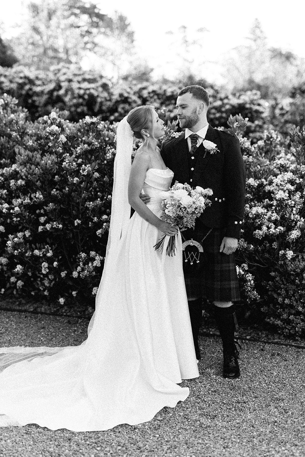 A black and white photo of a bride and groom standing together outdoors, surrounded by bushes and trees, sharing an intimate moment. The bride holds a bouquet of flowers, and the groom is dressed in traditional Scottish attire, including a kilt.