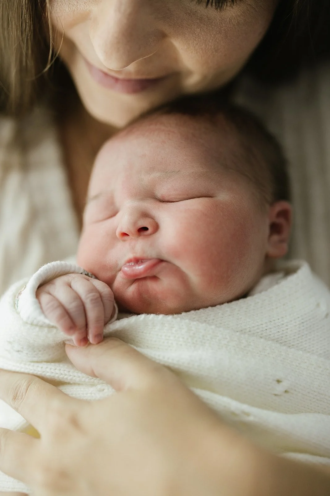 A close-up of a woman holding a sleeping newborn baby wrapped in a white blanket, with the baby's face resting against her chest.