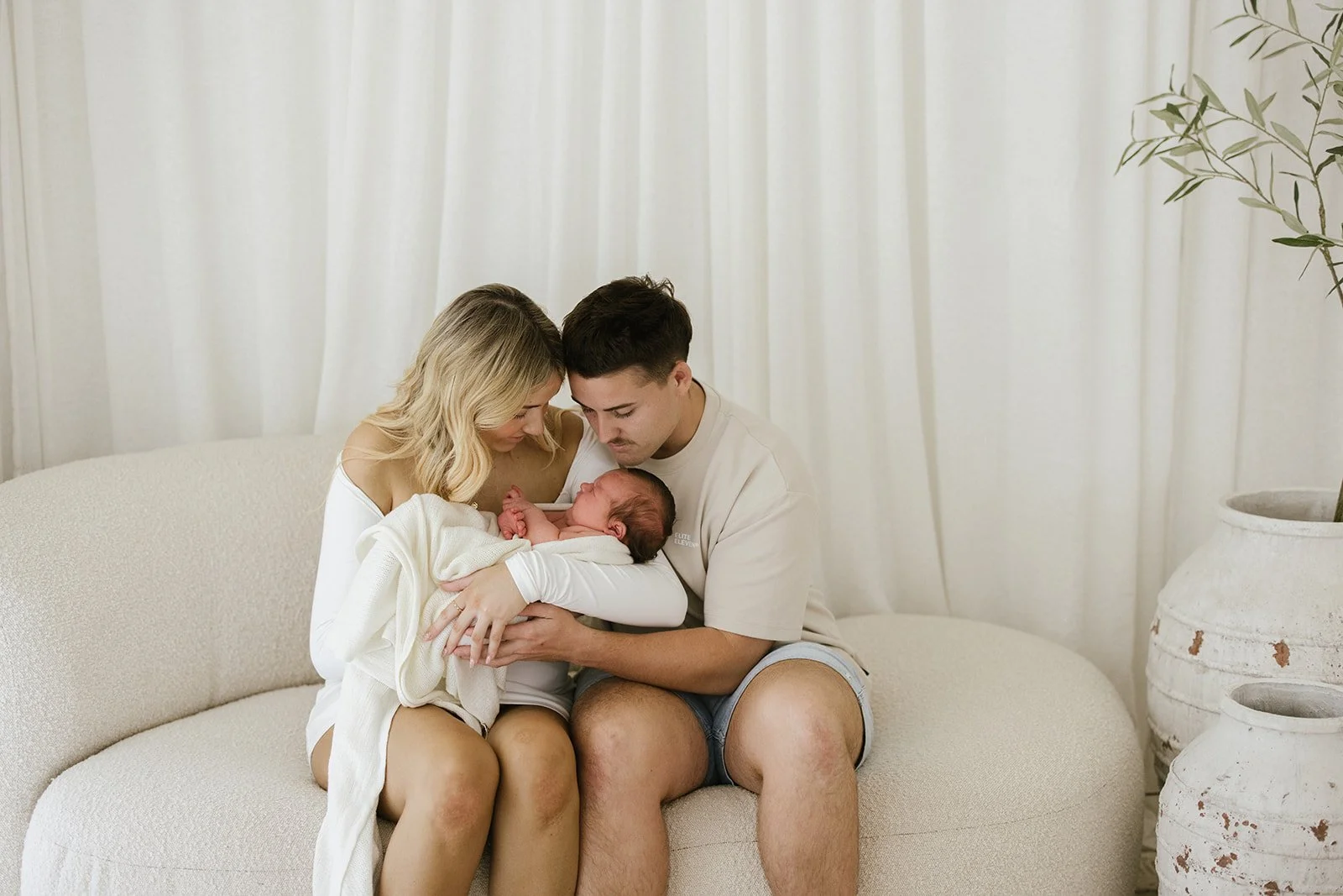 A family of three holding a newborn baby on a cream-colored sofa in a bright room with white curtains and large vases. Taken at Wylde Folk Studio - Brisbane newborn photographer.