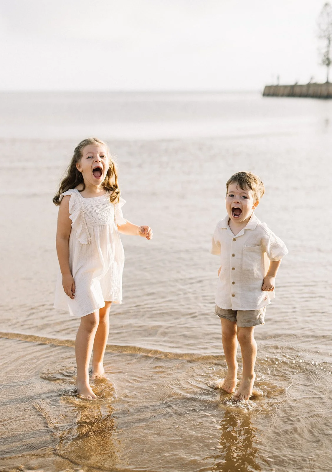 Two children, a girl and a boy, standing in shallow water at the beach, laughing and looking joyful, with a cloudy sky and a distant shoreline in the background. Taken by Wylde Folk Studio, Brisbane. Family photographer.