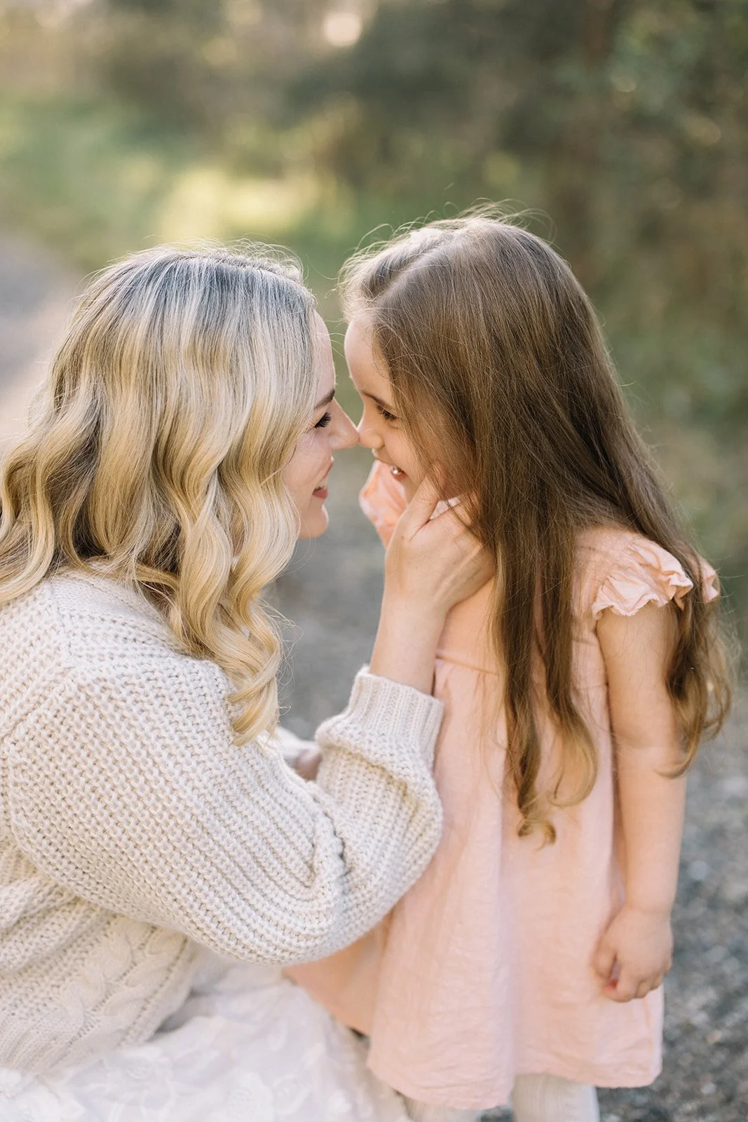A woman with blonde hair and a girl with brown hair touch noses and smile at each other outdoors. Taken by Wylde Folk Studio, Brisbane. Family photography.