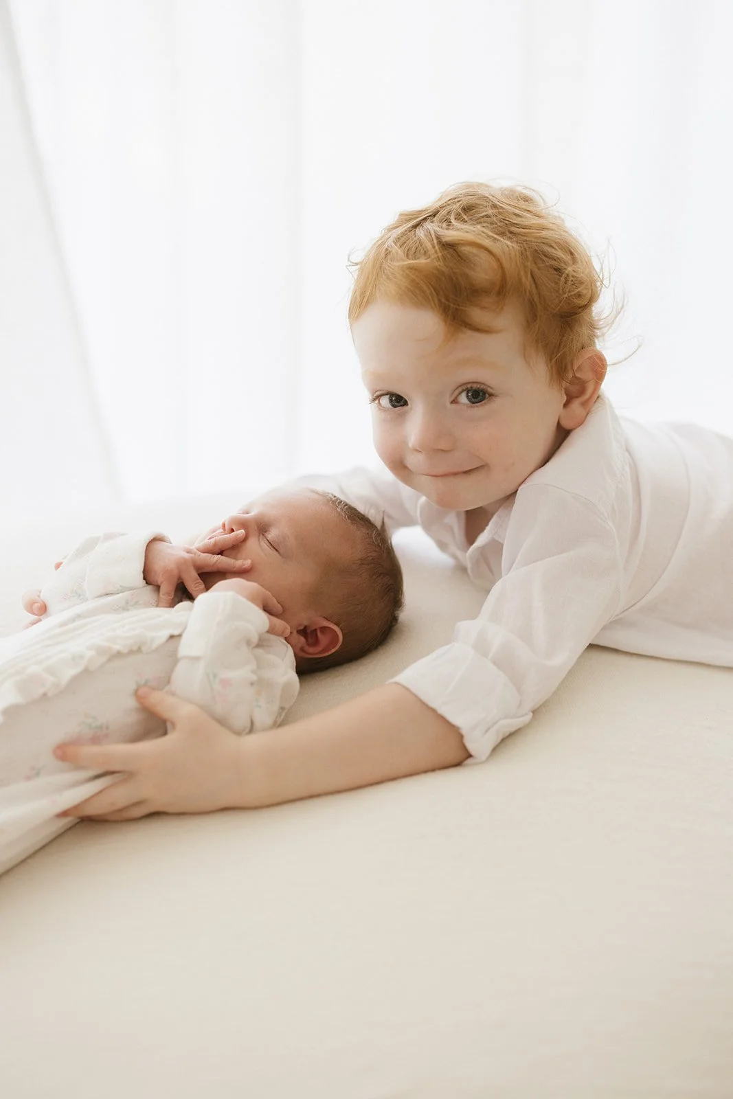 Newborn baby sleeps on white beanbag. Baby's brother with orange hair smiles at camera while having his arms around baby.
