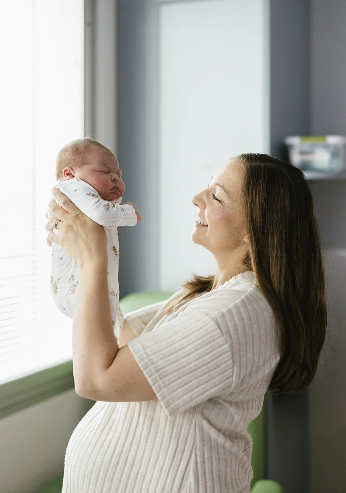 A woman holding a newborn baby and smiling, inside a room near a window.