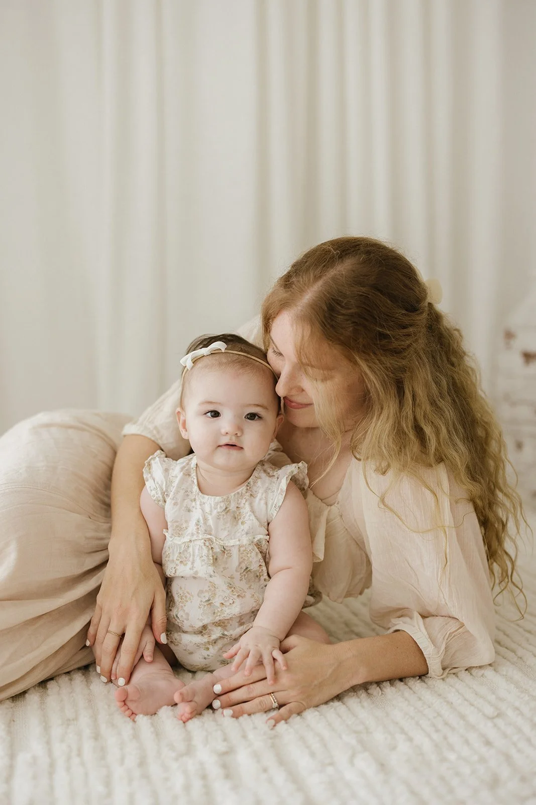 A woman with long, curly red hair lying on a cream-colored bed, with a young girl sitting on her lap. The girl has light brown hair with a bow and is wearing a floral pattern dress, while the woman is wearing a light-colored blouse. They are cuddling