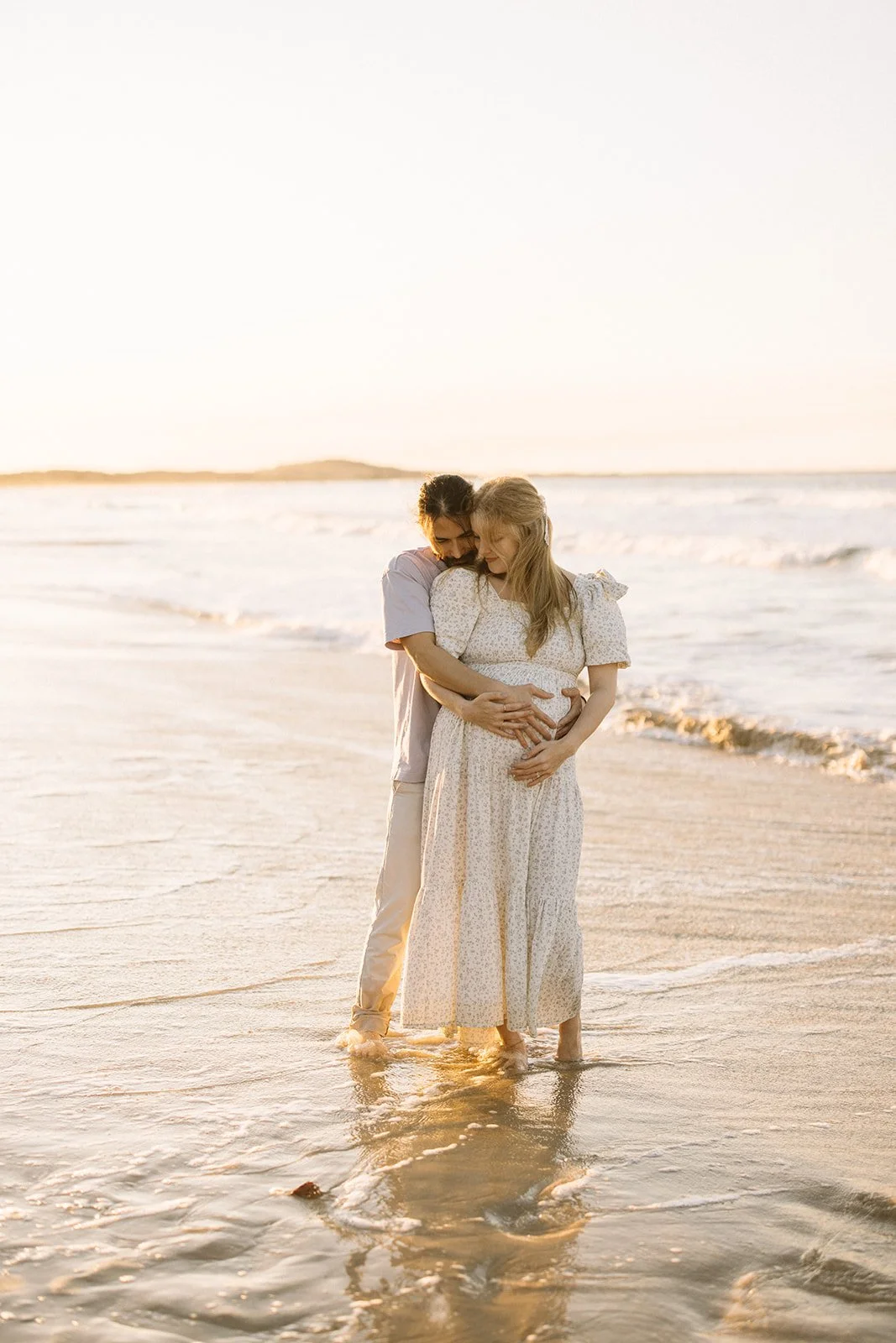 A couple standing in the shallow surf on a beach during sunset, embracing affectionately. Taken by Wylde Folk Studio, Brisbane. Maternity photographer.