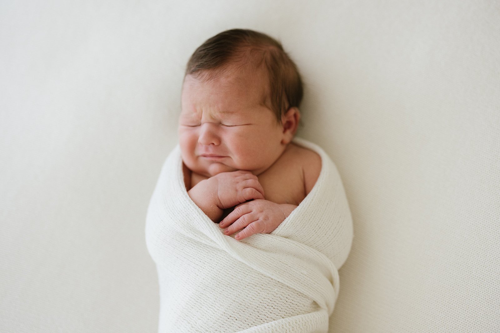 A newborn baby wrapped in a white knit blanket, sleeping with face scrunched and hands clasped near chest, against a plain white background.