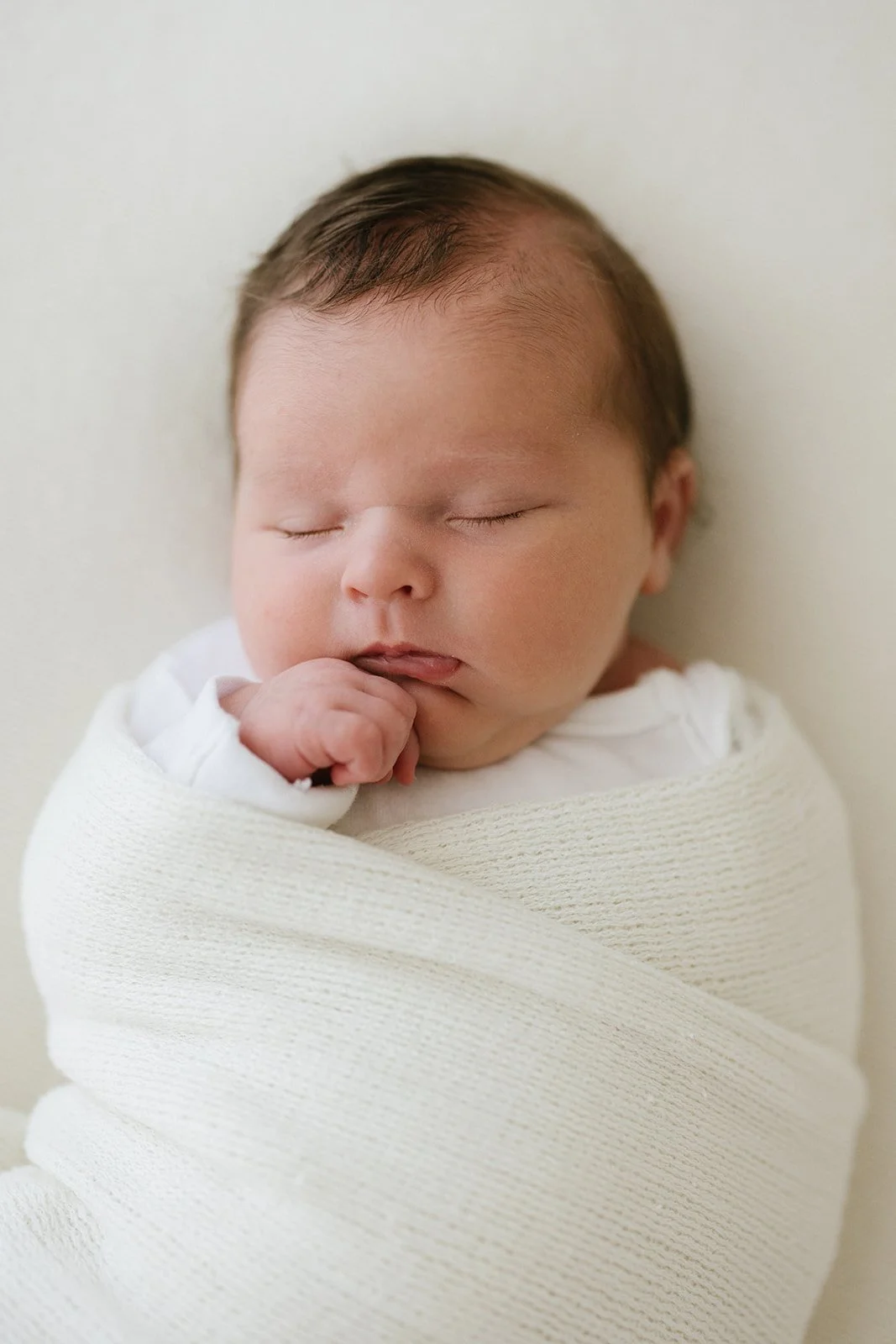 A sleeping newborn wrapped in a cream-colored blanket with a white shirt underneath, resting peacefully with a hand near the mouth.