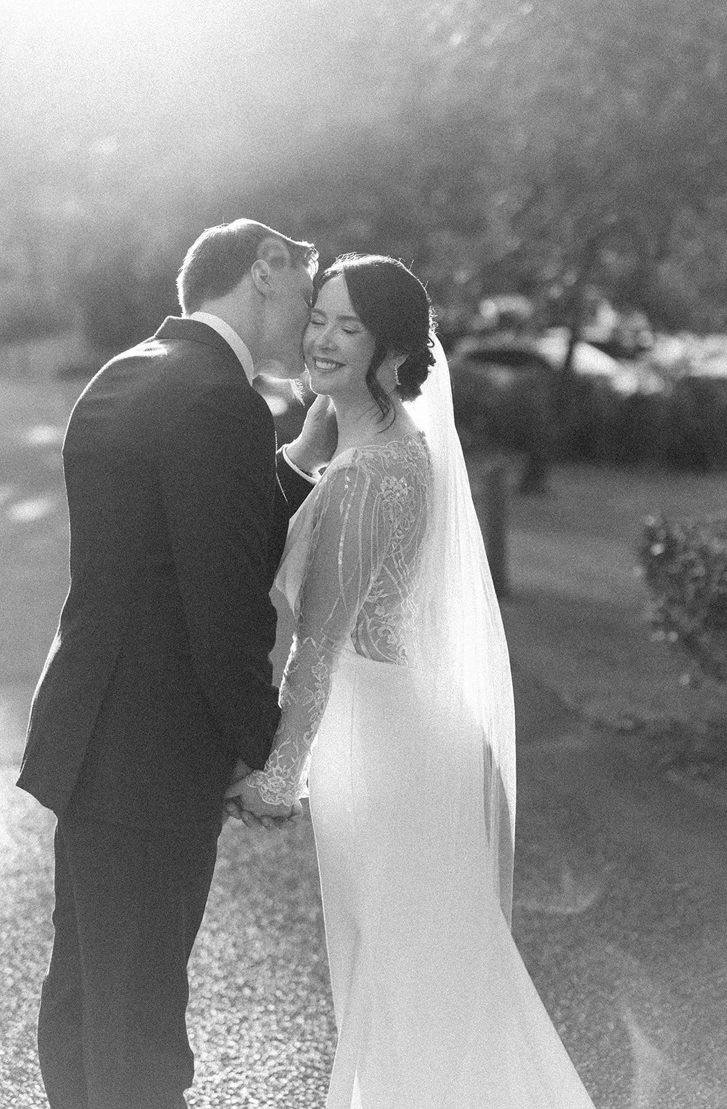 A black and white photo of a bride and groom on their wedding day, standing outdoors. The groom is kissing the bride's temple while holding her hand, and the bride is smiling with her eyes closed. The bride is wearing a lace wedding dress with long s