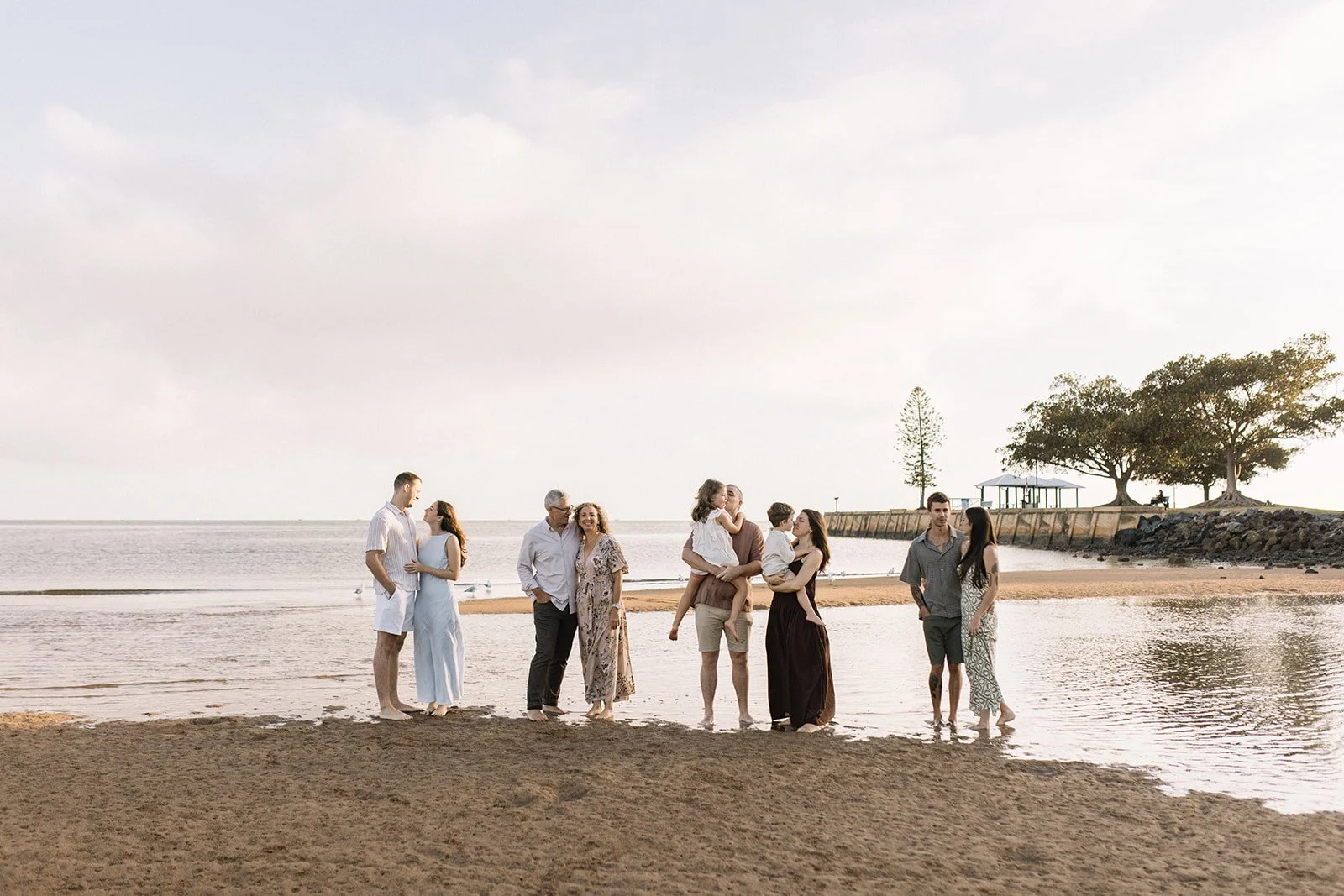 A group of eight people standing and walking on a beach near the water, with trees and a pavilion in the background during sunset or late afternoon. Taken by Wylde Folk Studio, Brisbane. Family photographer.