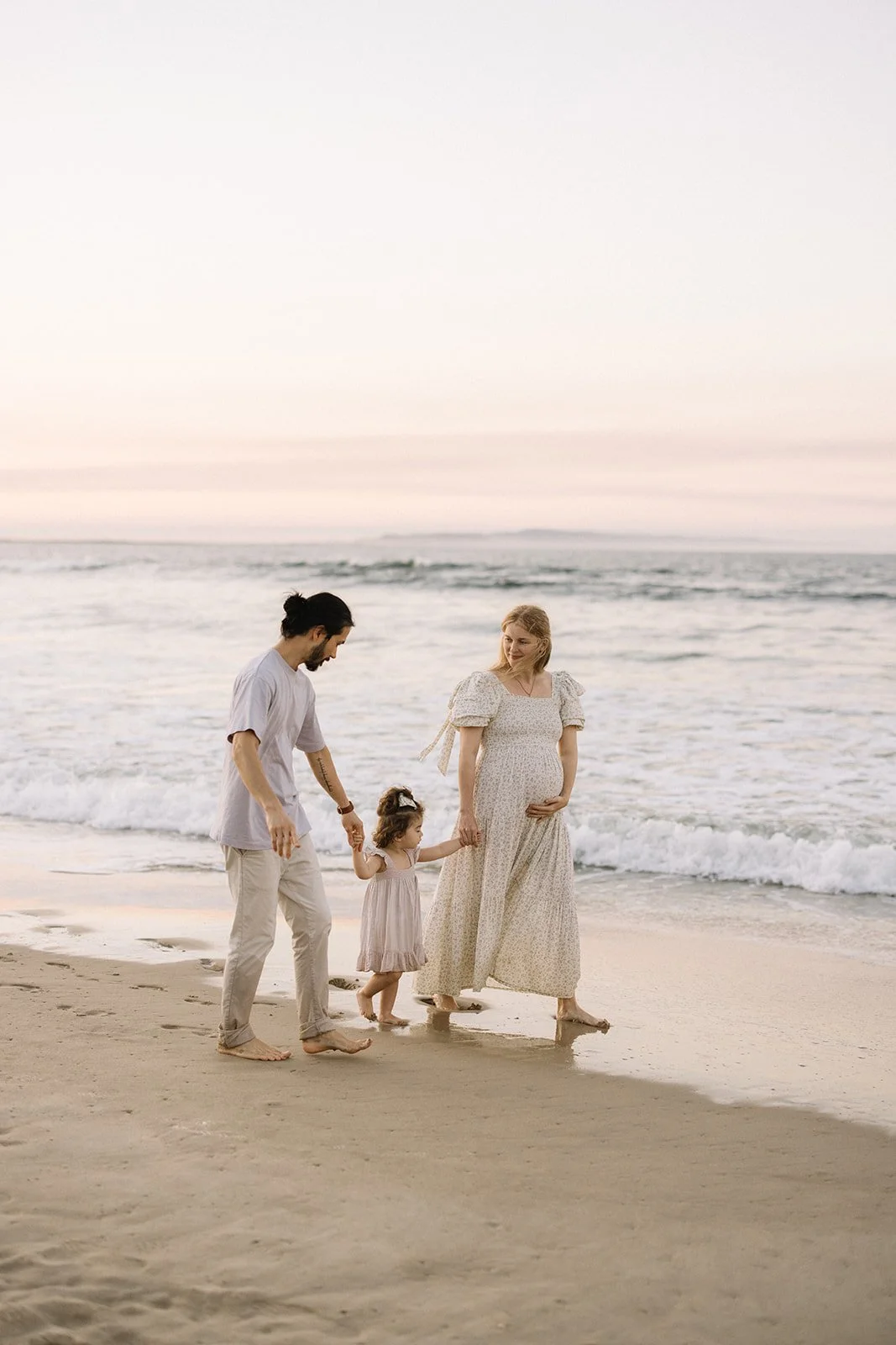 Family of three walking barefoot on the beach at sunset, holding hands, with the mother visibly pregnant. Taken by Wylde Folk Studio, Brisbane. Maternity photographer.