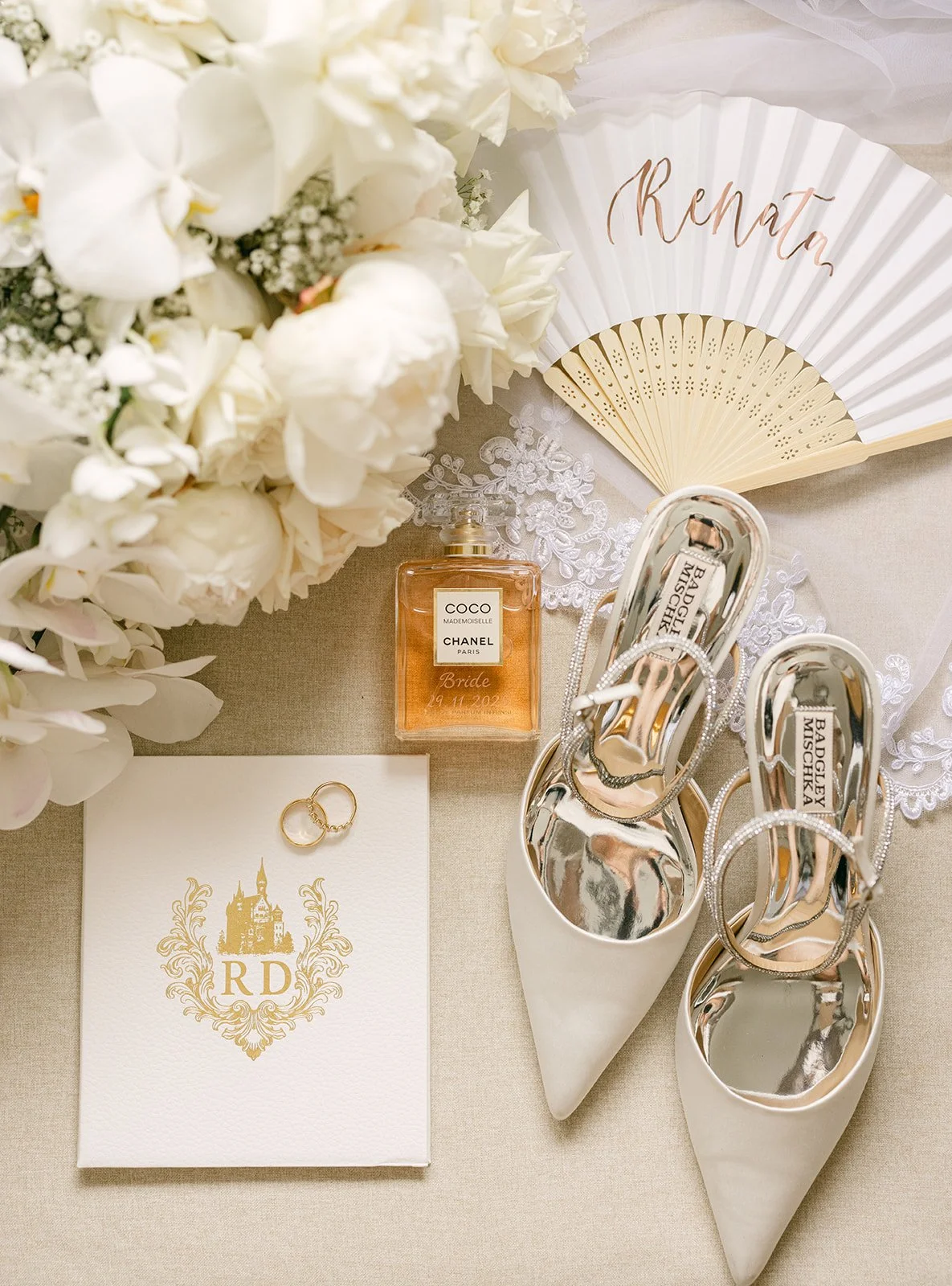 A wedding altar arrangement featuring a bouquet of white flowers, a fan with "Rena" written on it, a bottle of Chanel Coco Mademoiselle perfume labeled "Bride," a pair of white high heels with rhinestone ankle straps, a white card with gold crest and