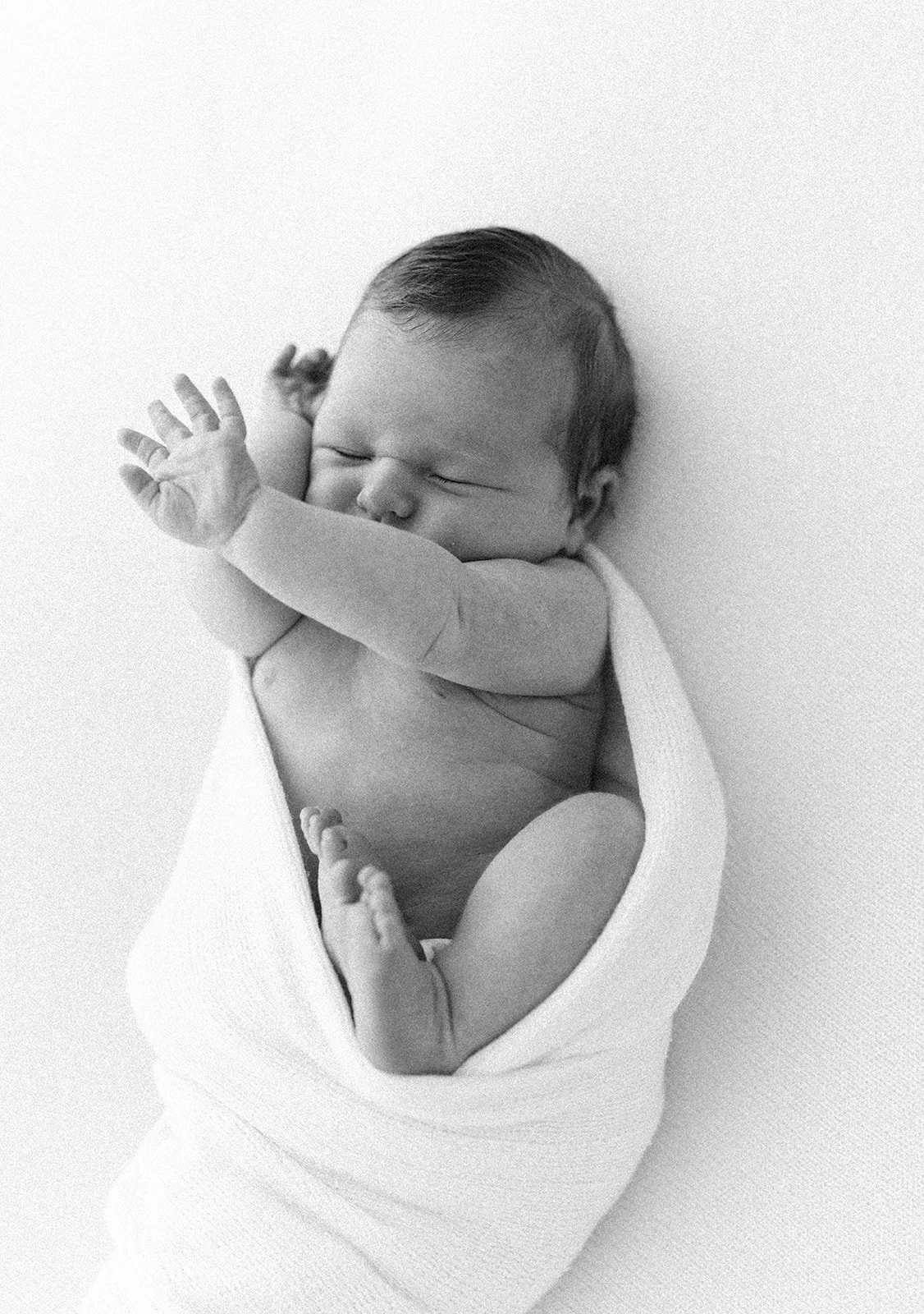 A black and white photo of a sleeping newborn baby wrapped in a white blanket, lying on a plain surface. Taken at Wylde Folk Studio - Brisbane newborn photographer.
