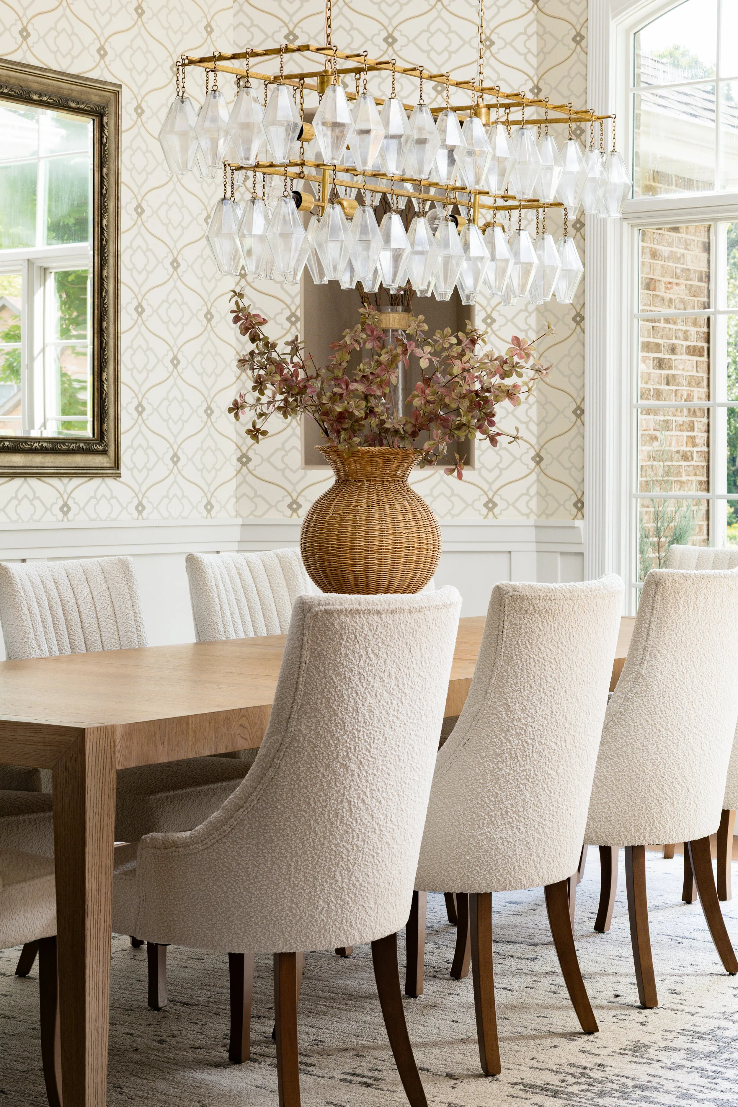 elegant and cozy dining room with wallpaper, a wood dining table with cream boucle dining chairs. The chandelier is gold with hanging crystals and a large-scale stem centerpiece. Southern-inspired interior design.