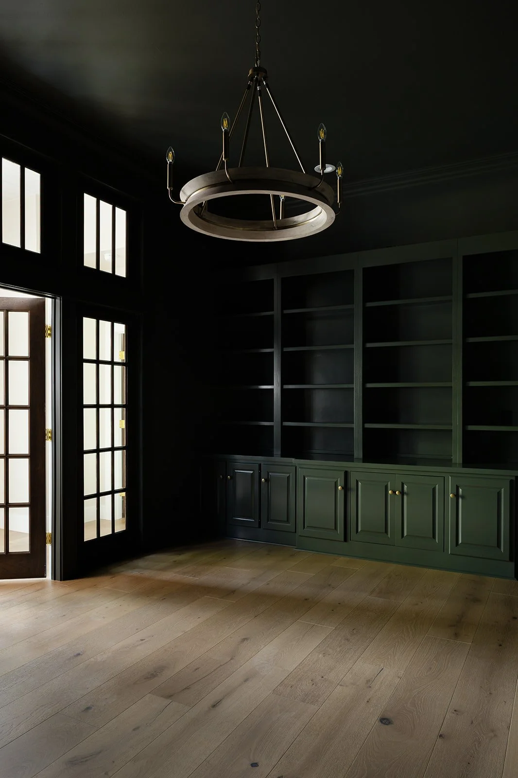 Empty room with black built-in shelves, green cabinet, wooden floor, black walls, and a black chandelier hanging from the ceiling.