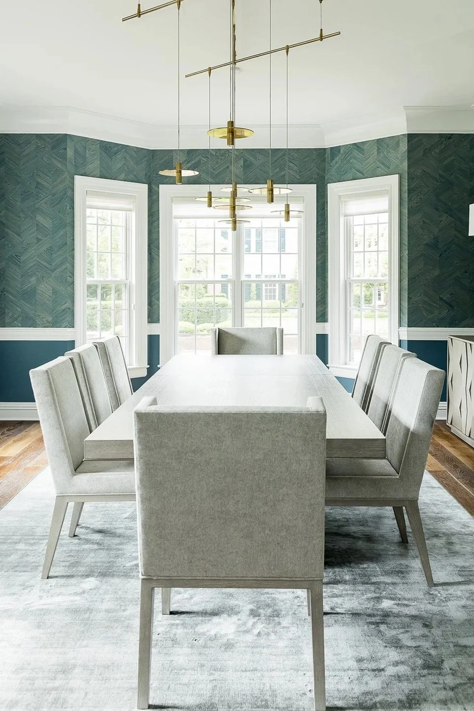 Modern dining room with a white table, grey upholstered chairs, and large windows, with a brass chandelier overhead.