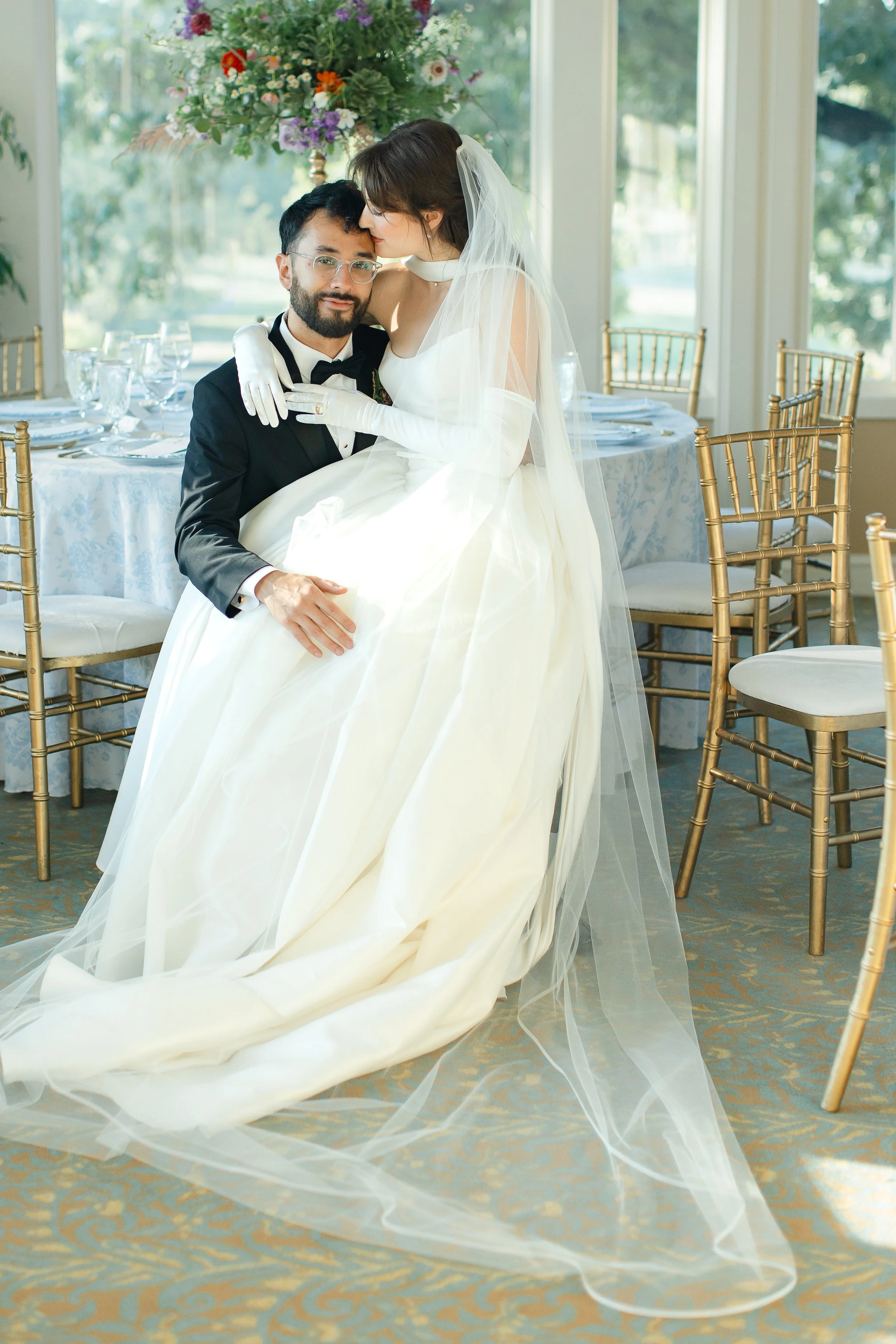A bride and groom at their wedding reception, with the bride kissing the groom on the forehead while he sits and looks at the camera. The bride is wearing a white wedding gown with a long veil and white gloves, and the groom is wearing a black tuxedo