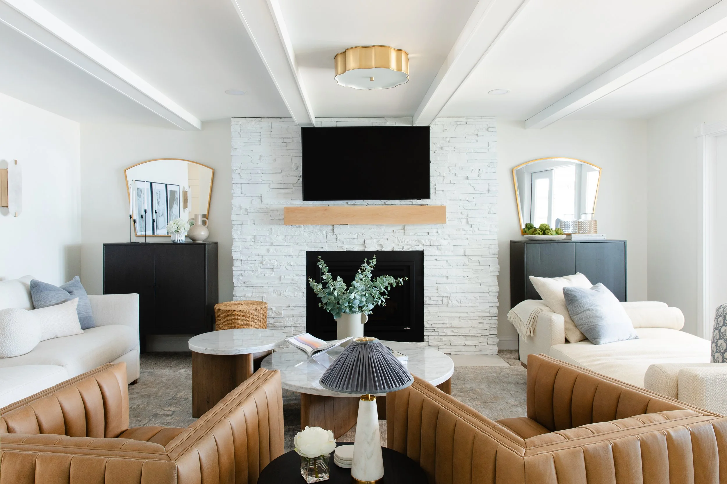 A modern living room featuring a white brick fireplace with a black fireplace insert, surrounded by comfortable sofas and armchairs in neutral tones. There are two black cabinets with decorative mirrors and vases on top, and a coffee table with a dec