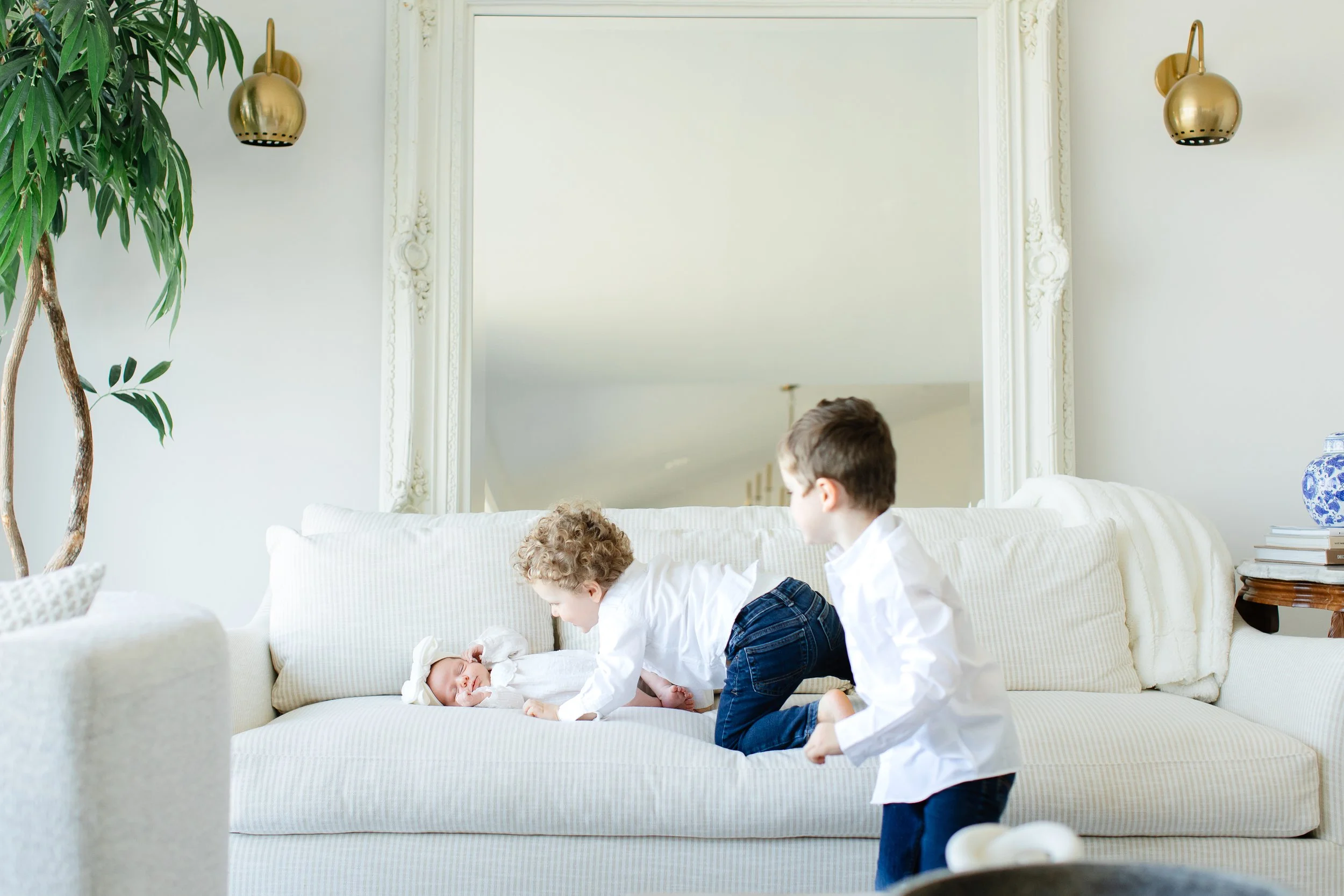 Three children on a white couch, two boys and one baby, with a mirror, a plant, and decorative lamp in a bright living room.