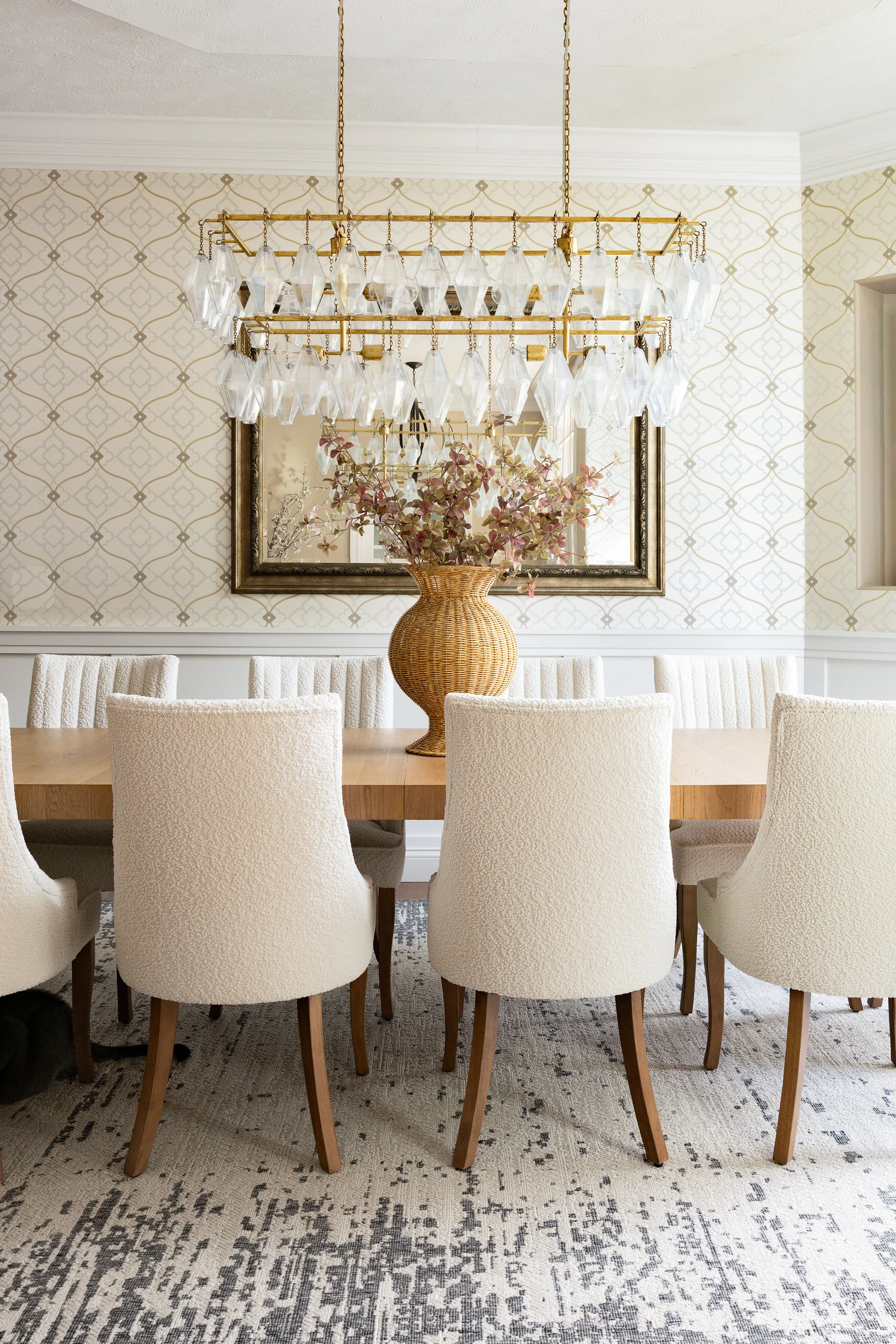 Wallpapered dining room with a big gold and crystal chandelier over a wood table with boucle, cream dining chairs. Southern inspired interior design.
