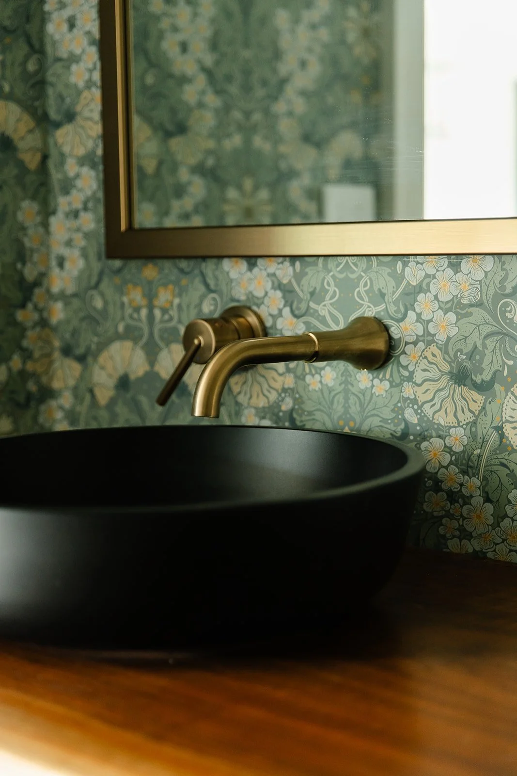 Close-up of a black vessel sink with a brass wall-mounted faucet above it, against a floral wallpaper background with a mirror above.