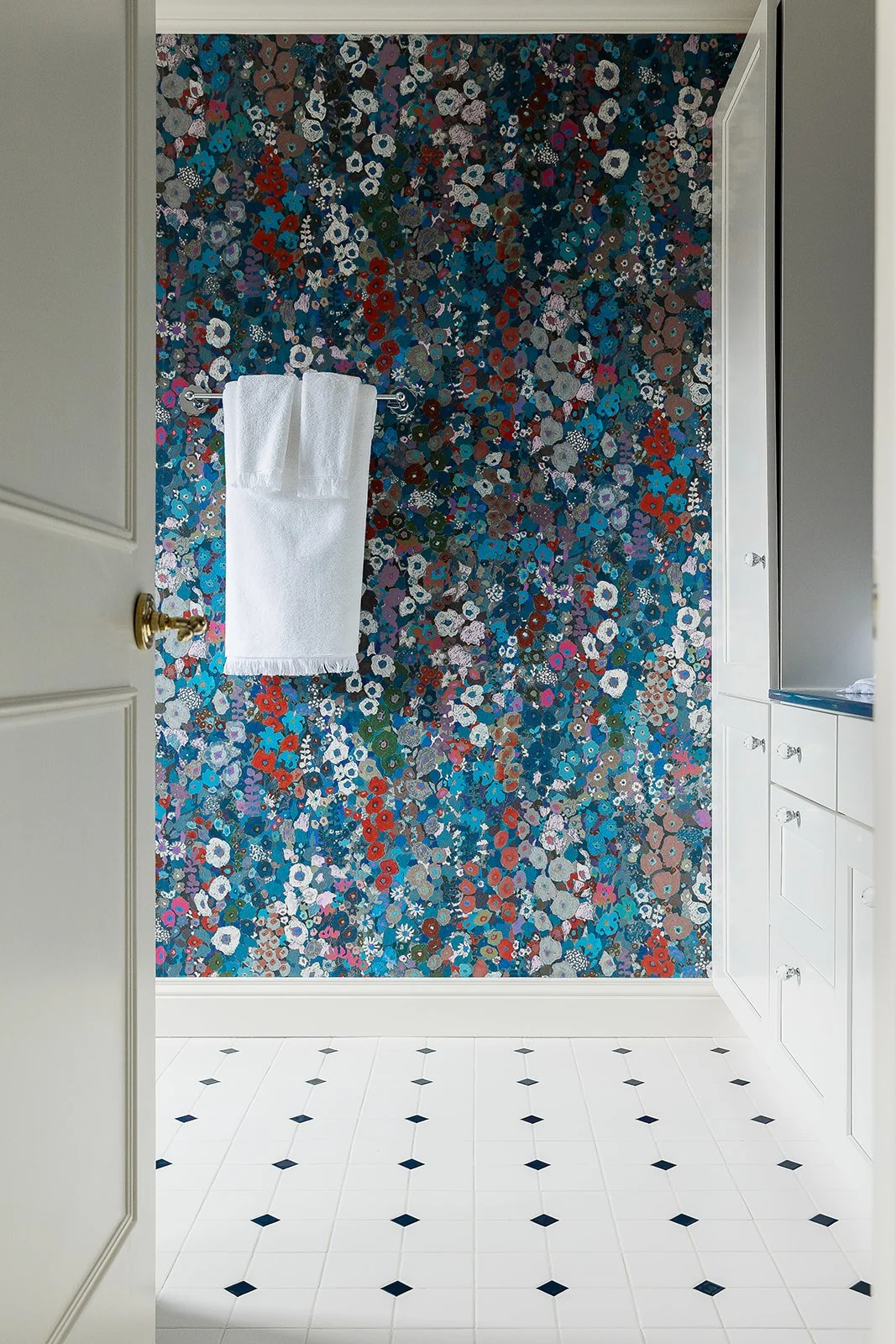 A bathroom with a floral patterned wallpaper wall, white tile floor with small black diamond accents, a towel rack with white towels, and white cabinetry.